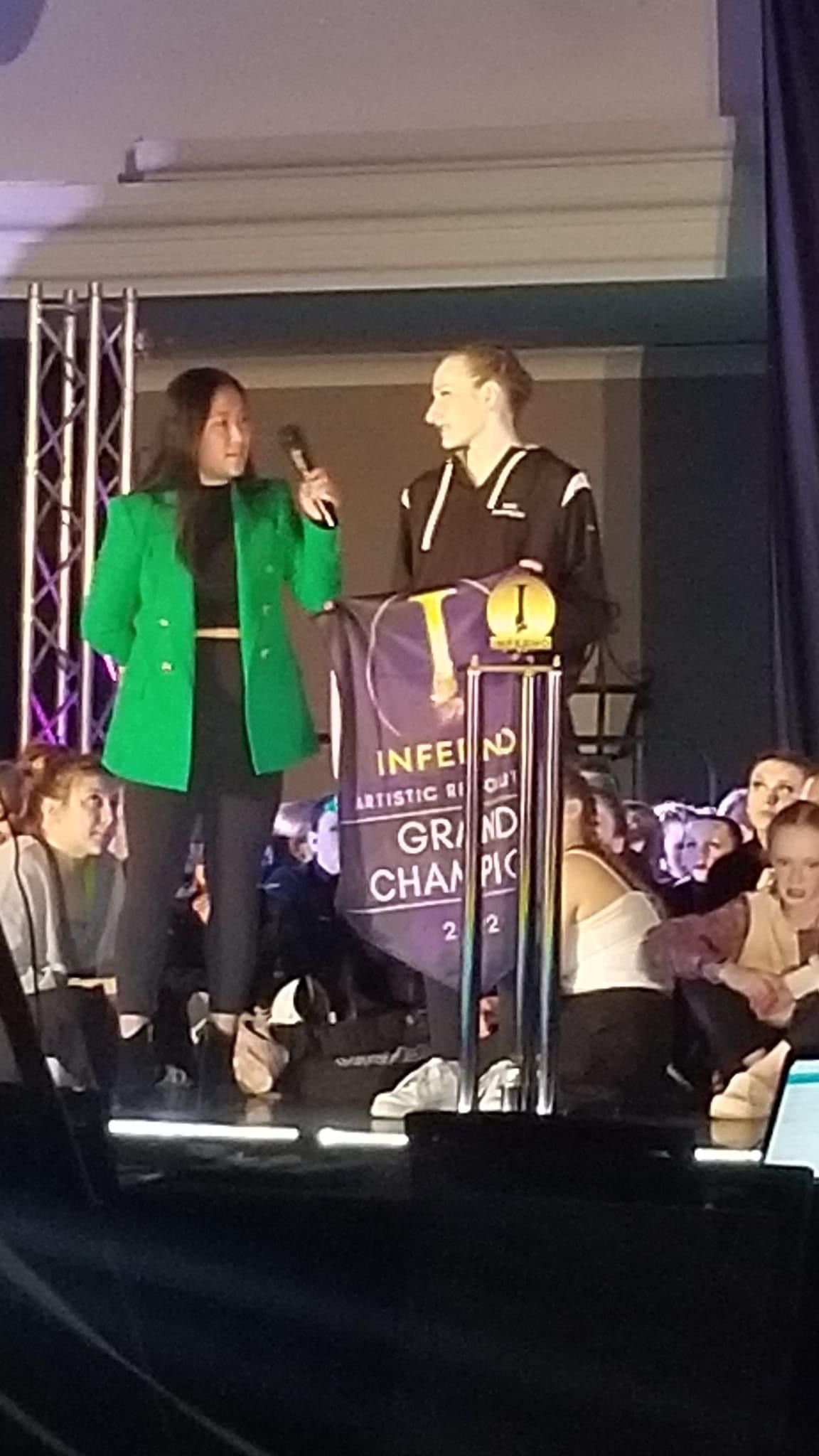 A woman in a green jacket is standing on a stage holding a microphone