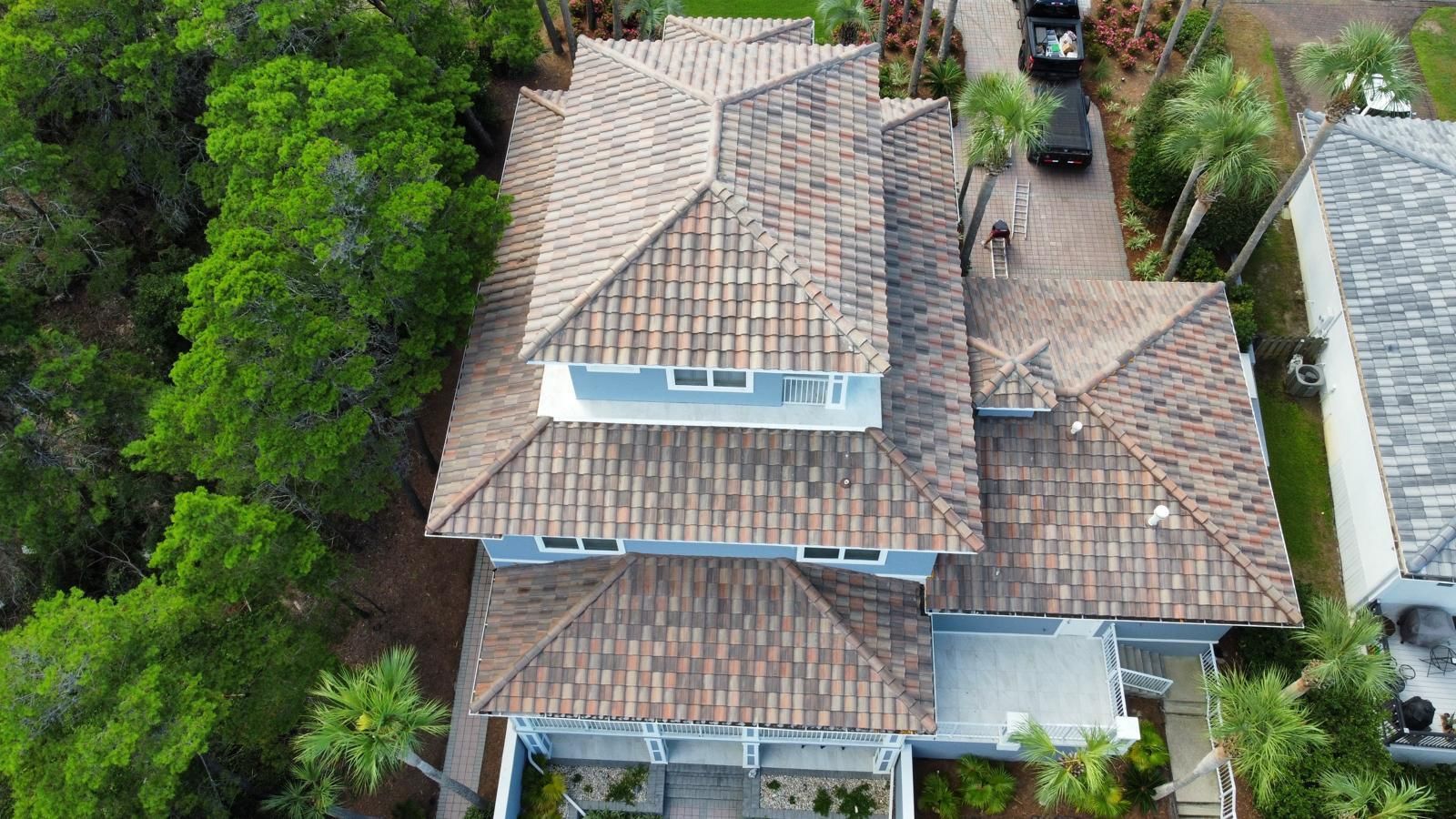 Aerial view of a blue house with a brown tile roof and driveway, surrounded by trees.