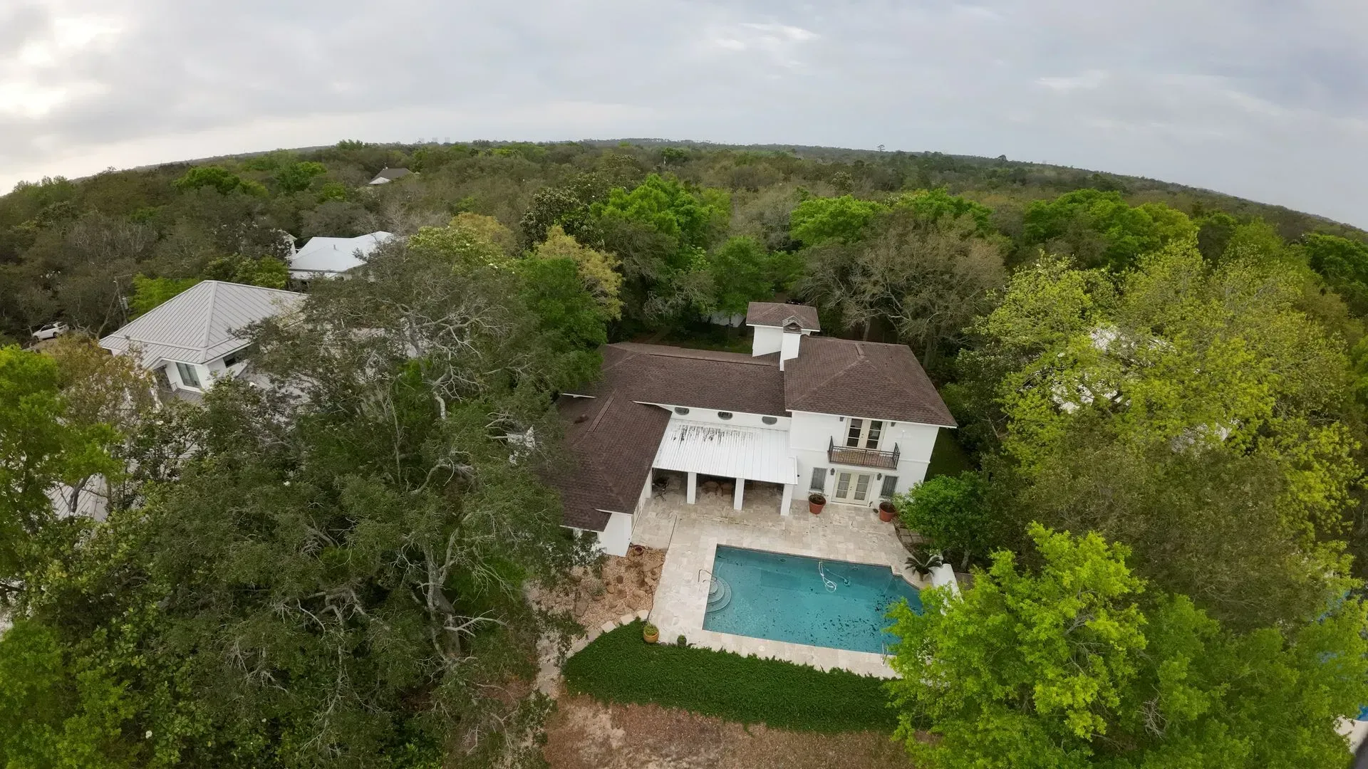 Aerial view of a white house with a pool surrounded by trees, under a cloudy sky.