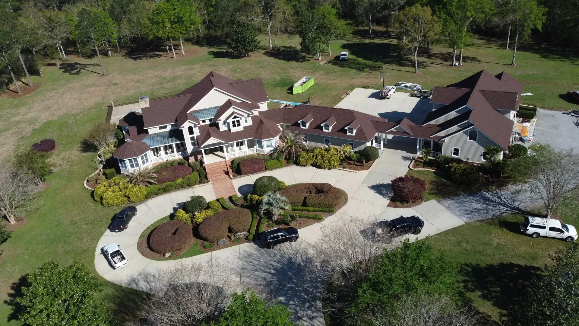 Aerial view of a large house with a circular driveway and several cars parked in front, surrounded by trees and greenery.