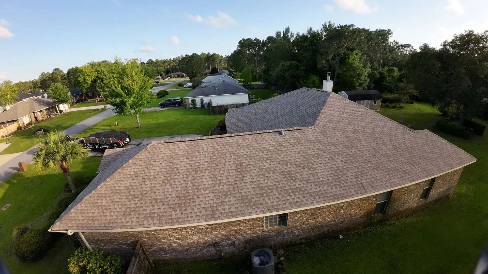 Aerial view of a house with a brown roof, green grass, and surrounding trees on a sunny day.
