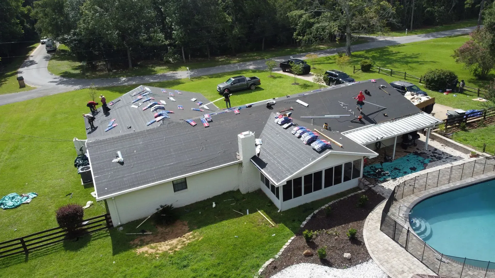 Roofers working on a dark-shingled roof of a white house with a pool.