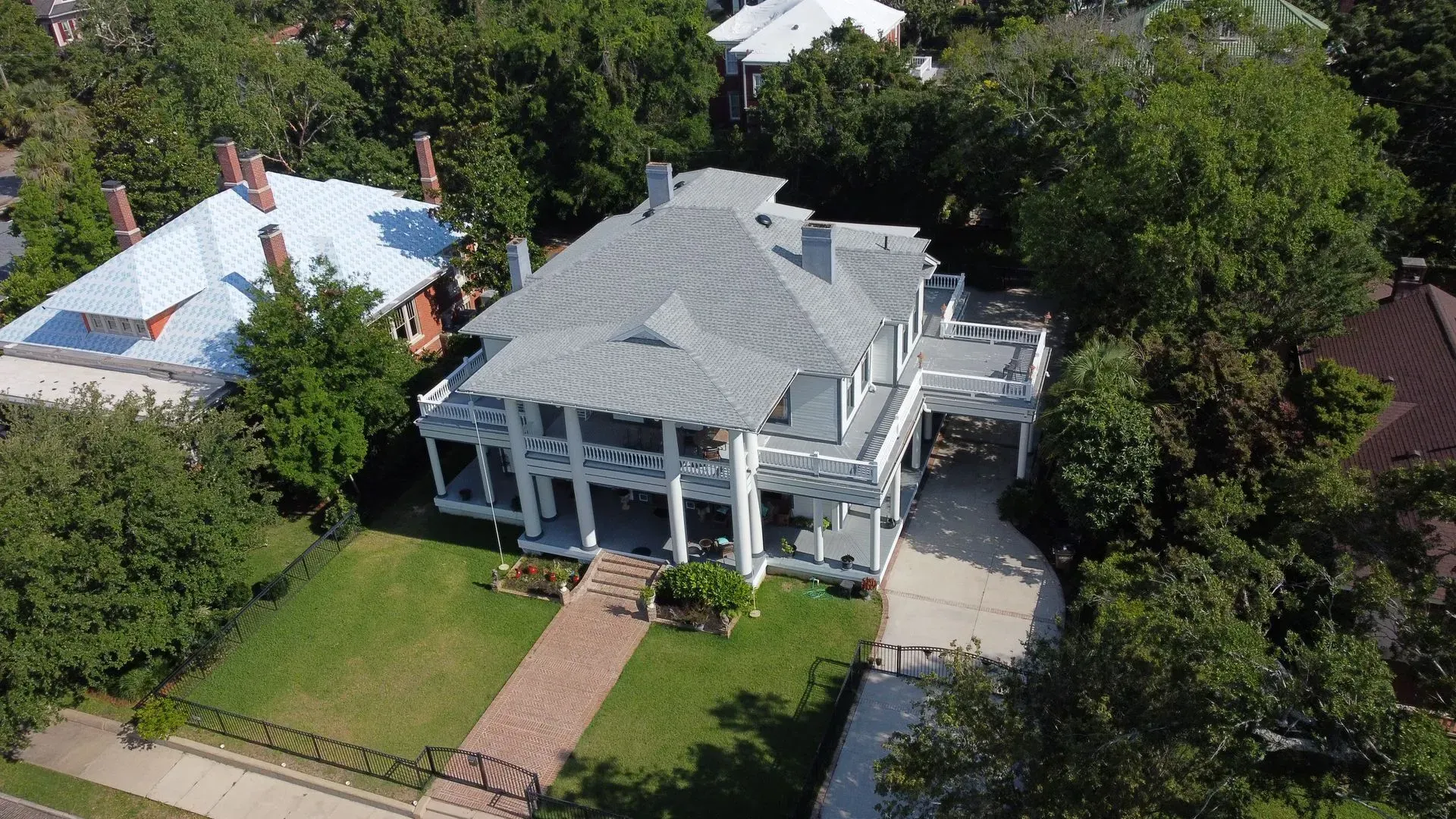White two-story house with wrap-around porch, brick walkway, and green lawn, nestled amongst trees.