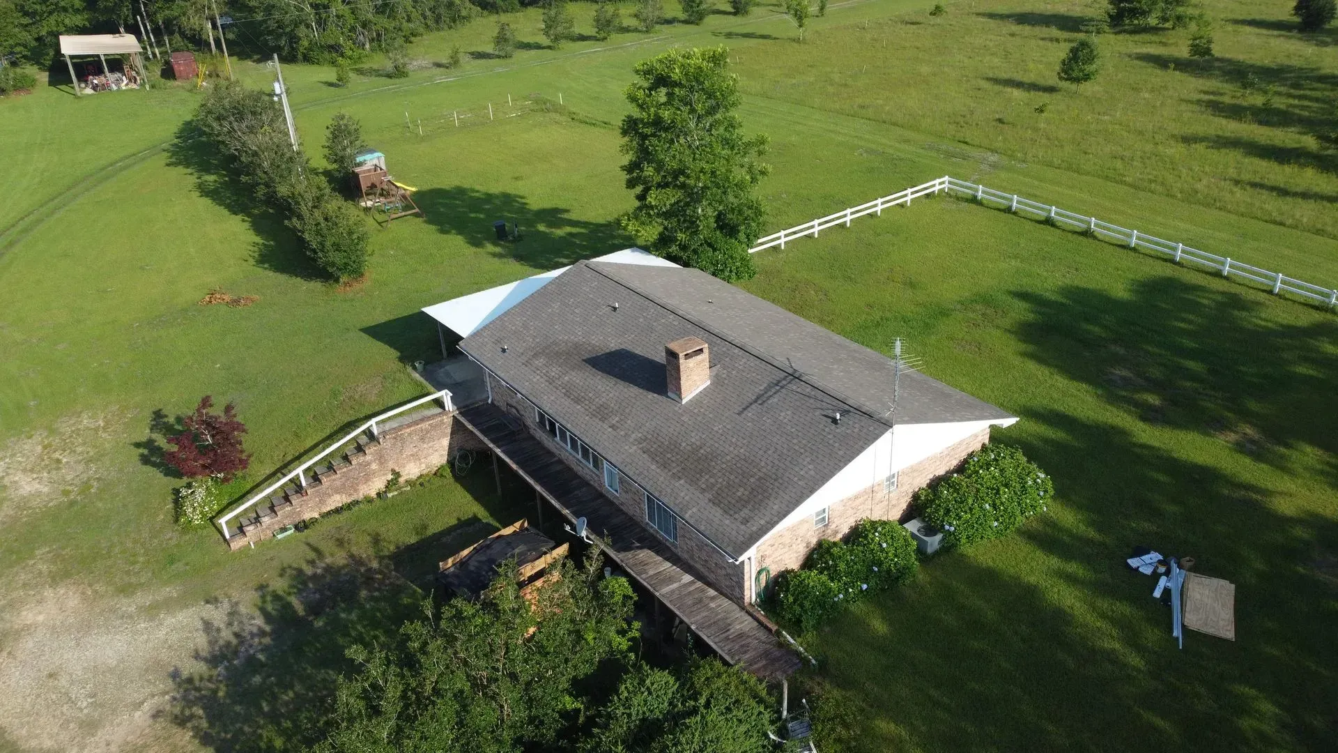 Aerial view of a house with a gray roof on a large green lawn, surrounded by trees and a white fence.