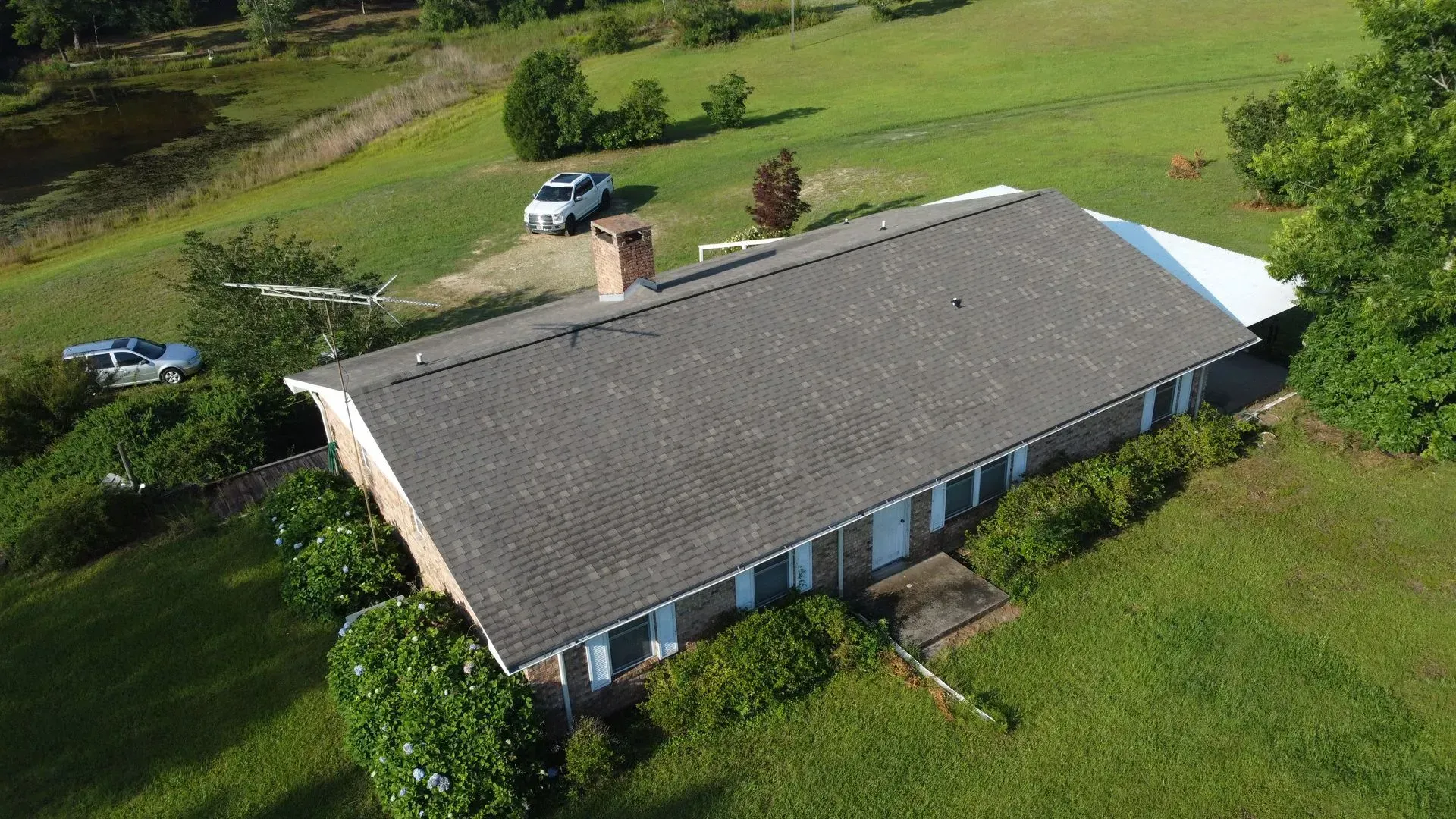Aerial view of a single-story house with a brick chimney and white trim, surrounded by green grass and trees.