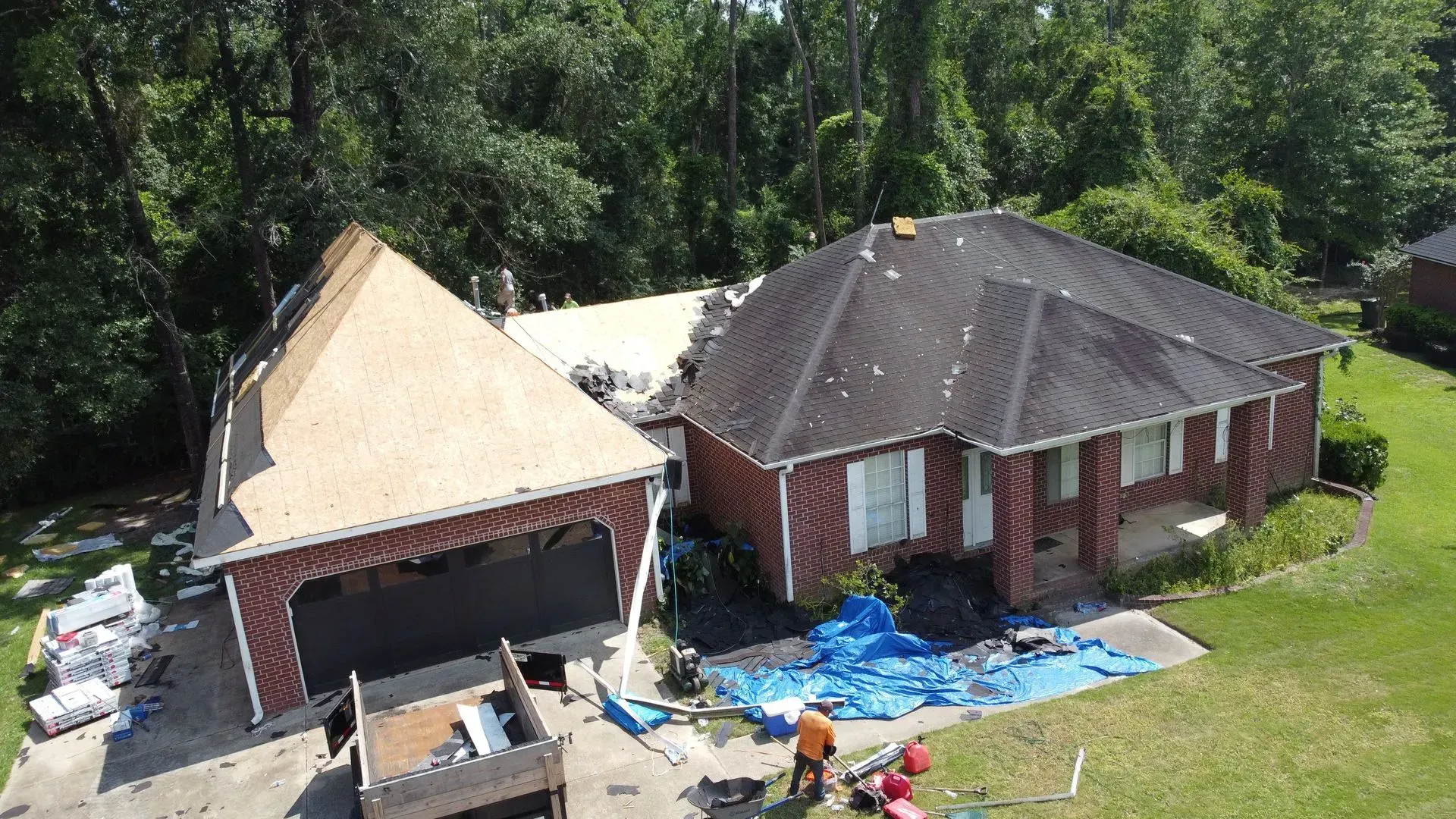 House with roof damage; workers on the ground and partially covered with a blue tarp. Green trees in the background.