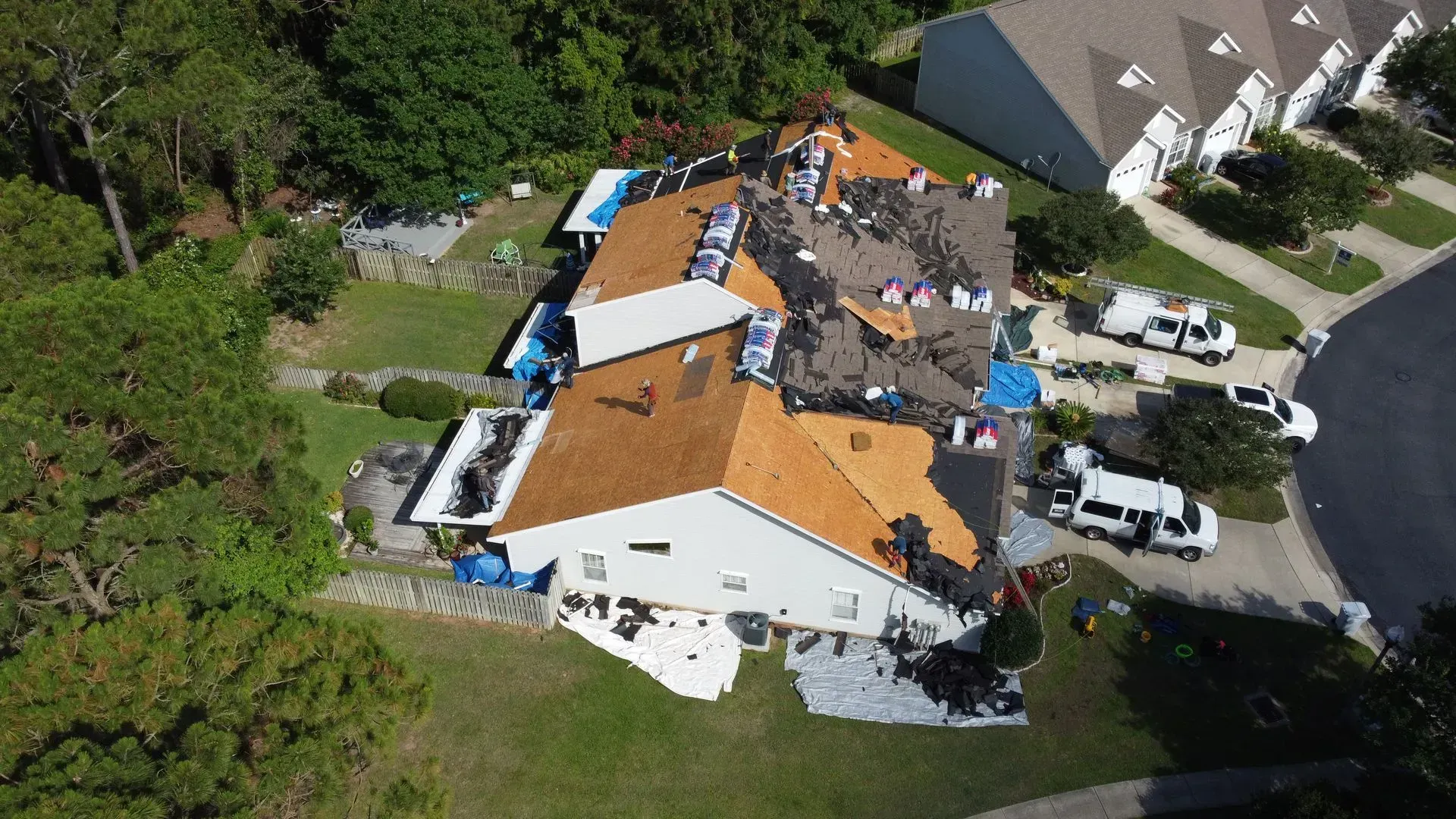 Aerial view of a house undergoing roof replacement, with multiple work trucks parked nearby.