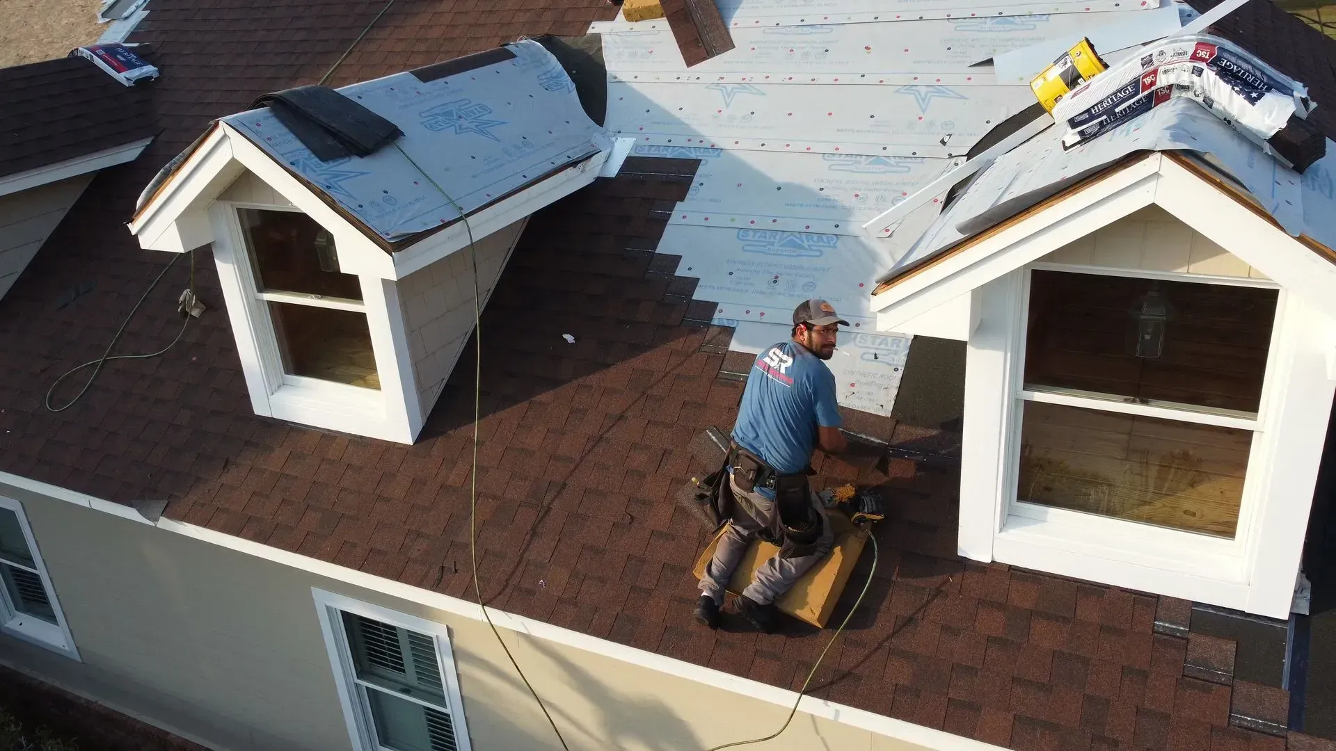 Roofer on a brown-shingled roof, near dormers, working. Wearing tool belt, blue shirt. Sunny day.