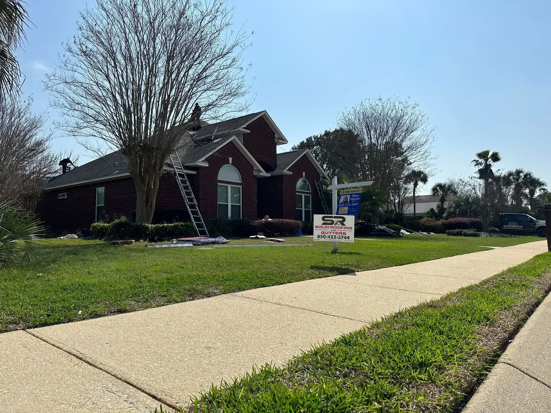House with a brick facade, ladder on the roof, sign in the yard, and a green lawn under a sunny sky.