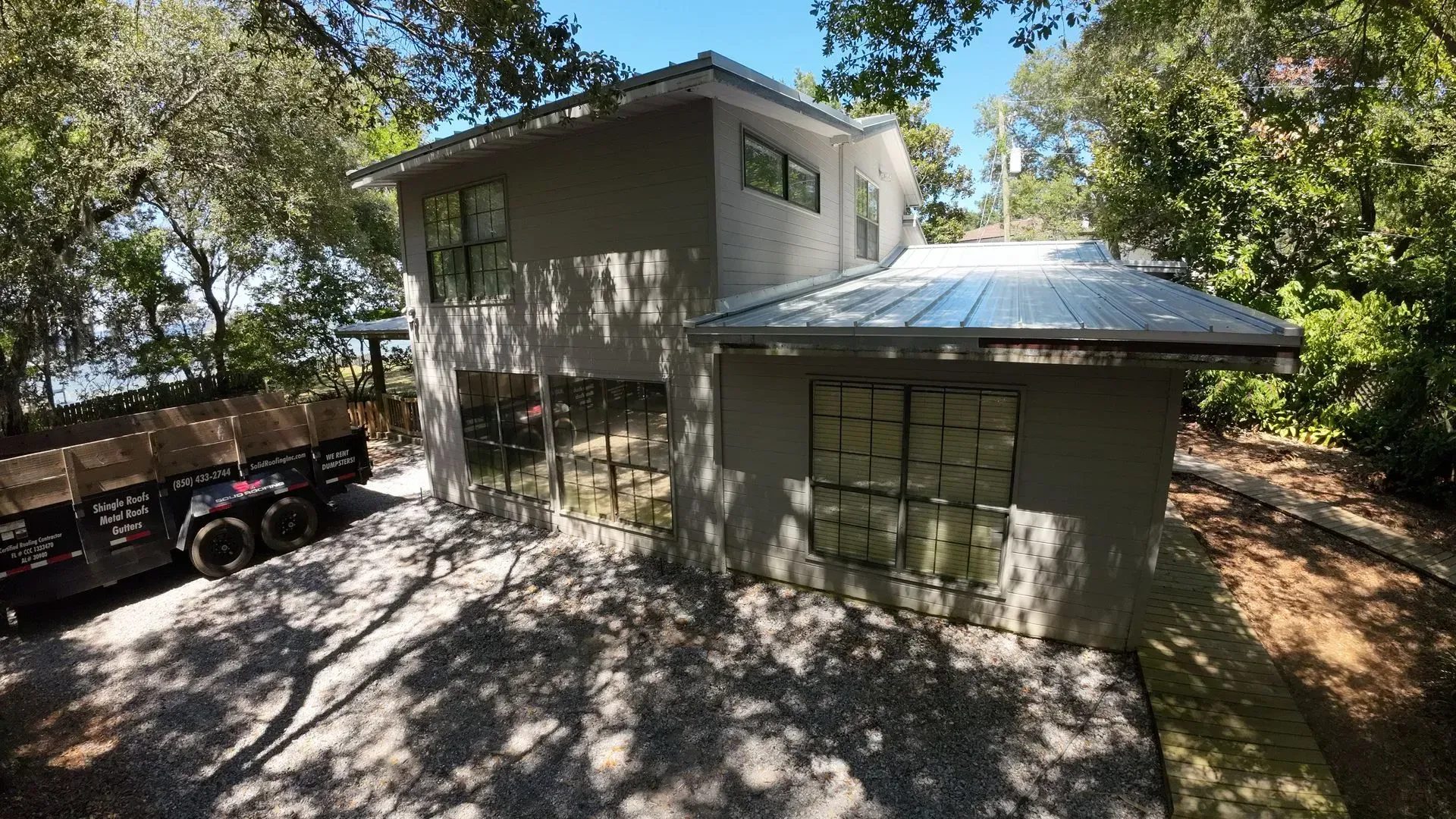 Two-story modern house with a gray exterior and gravel driveway, nestled among trees, and a trailer parked nearby.