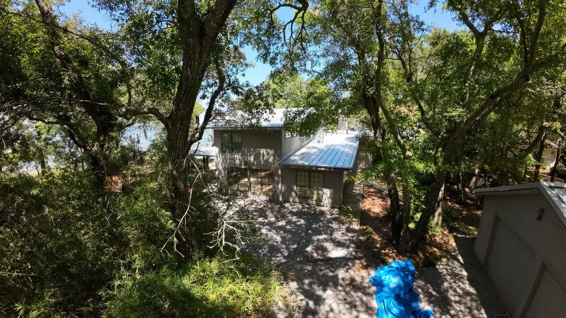 A house is nestled among green trees. The roof is silver, and a blue tarp lies on the ground nearby.