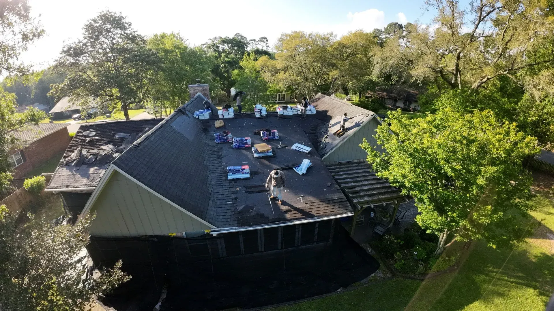 Workers on a roof removing old shingles, surrounded by trees.