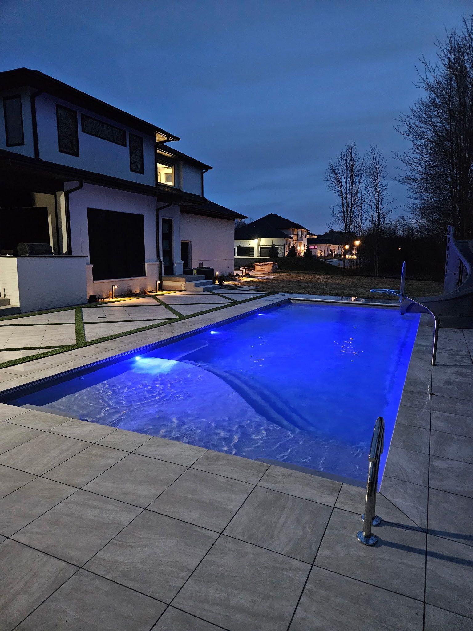 Blue-lit rectangular pool beside a white house at dusk. Concrete patio, stainless steel handrails.