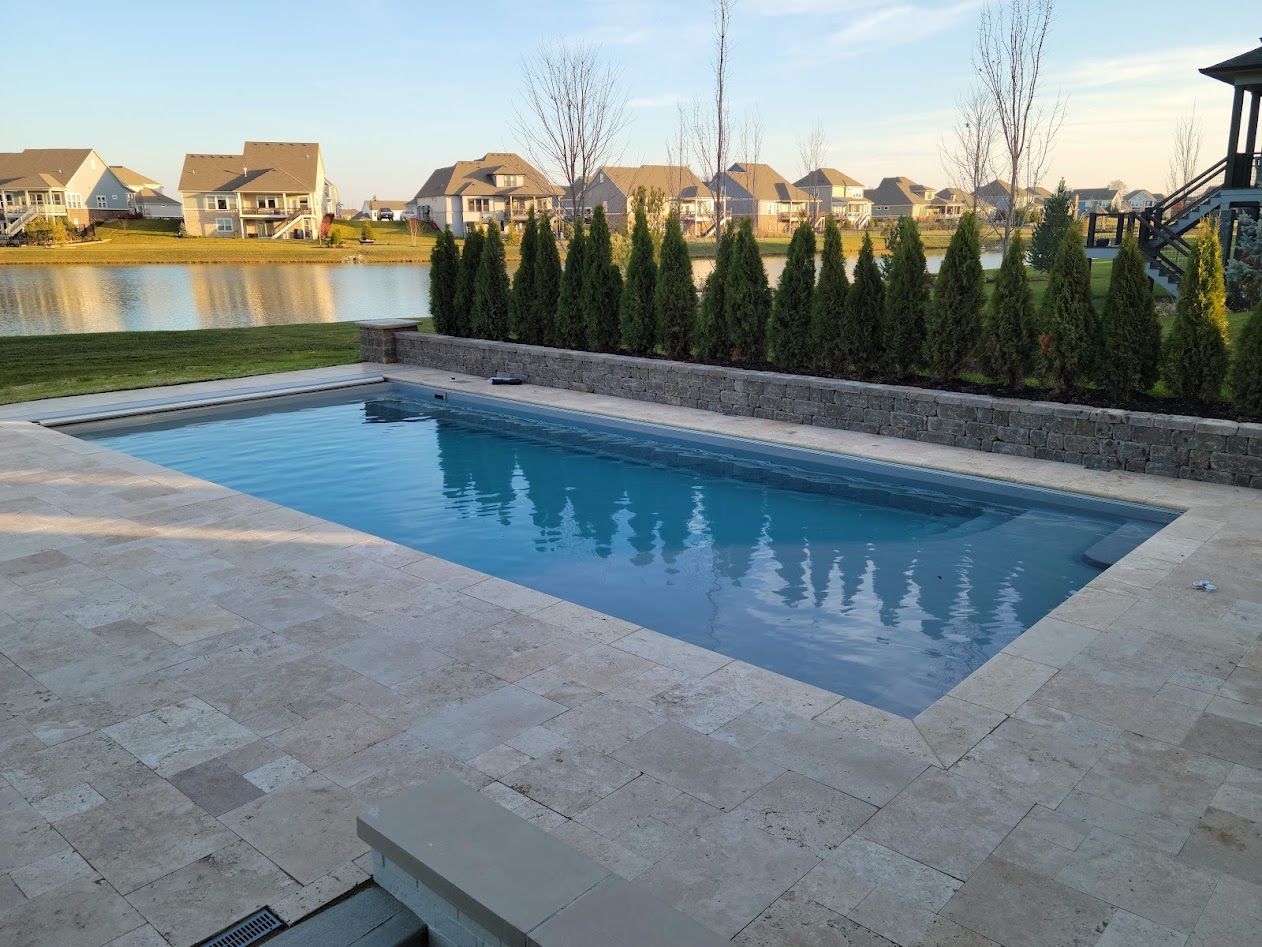 Rectangular pool with travertine patio, backed by tall green shrubs, and suburban homes.