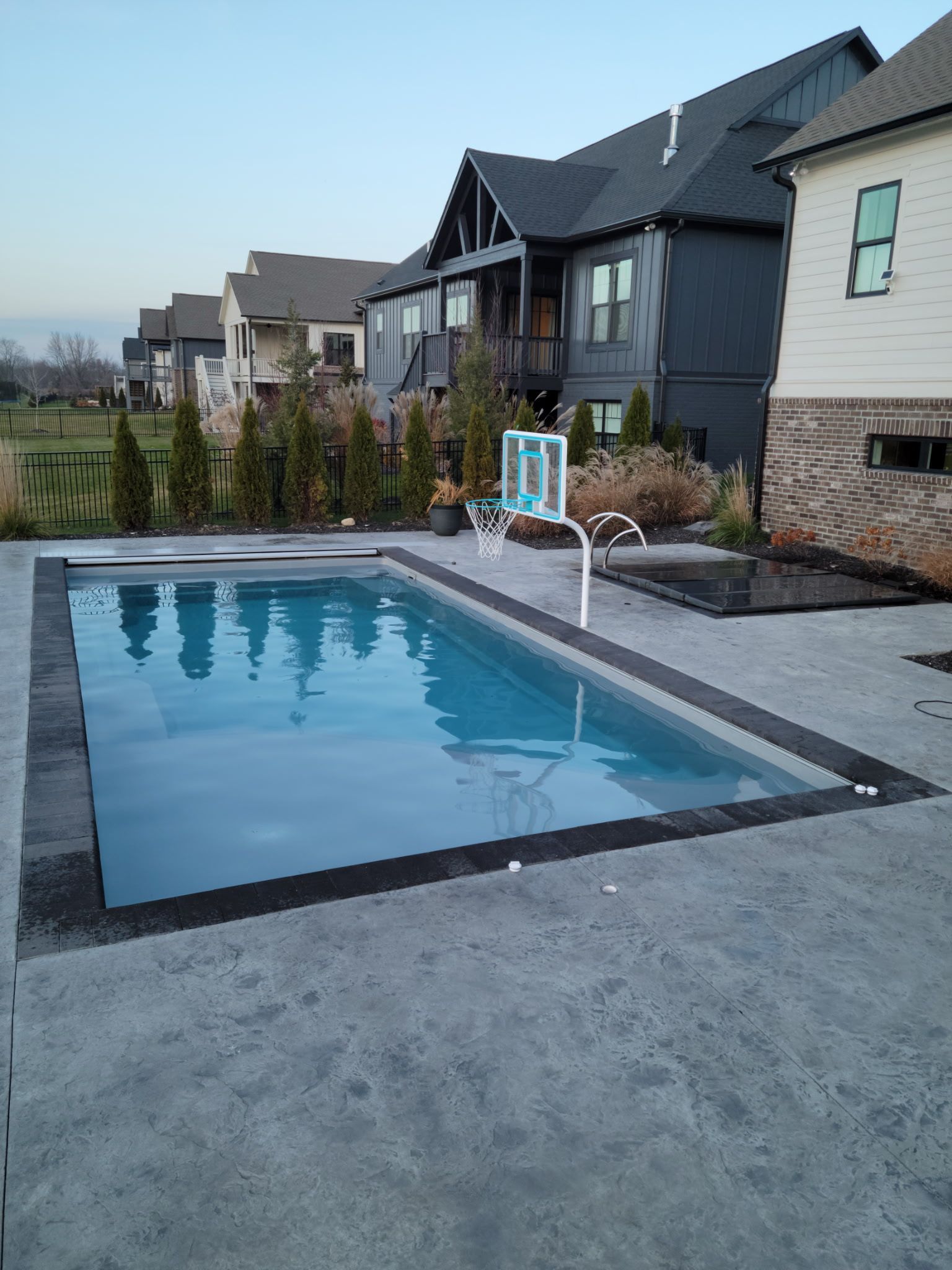 Swimming pool with basketball hoop in backyard, houses in the background. Blue water, gray concrete patio.