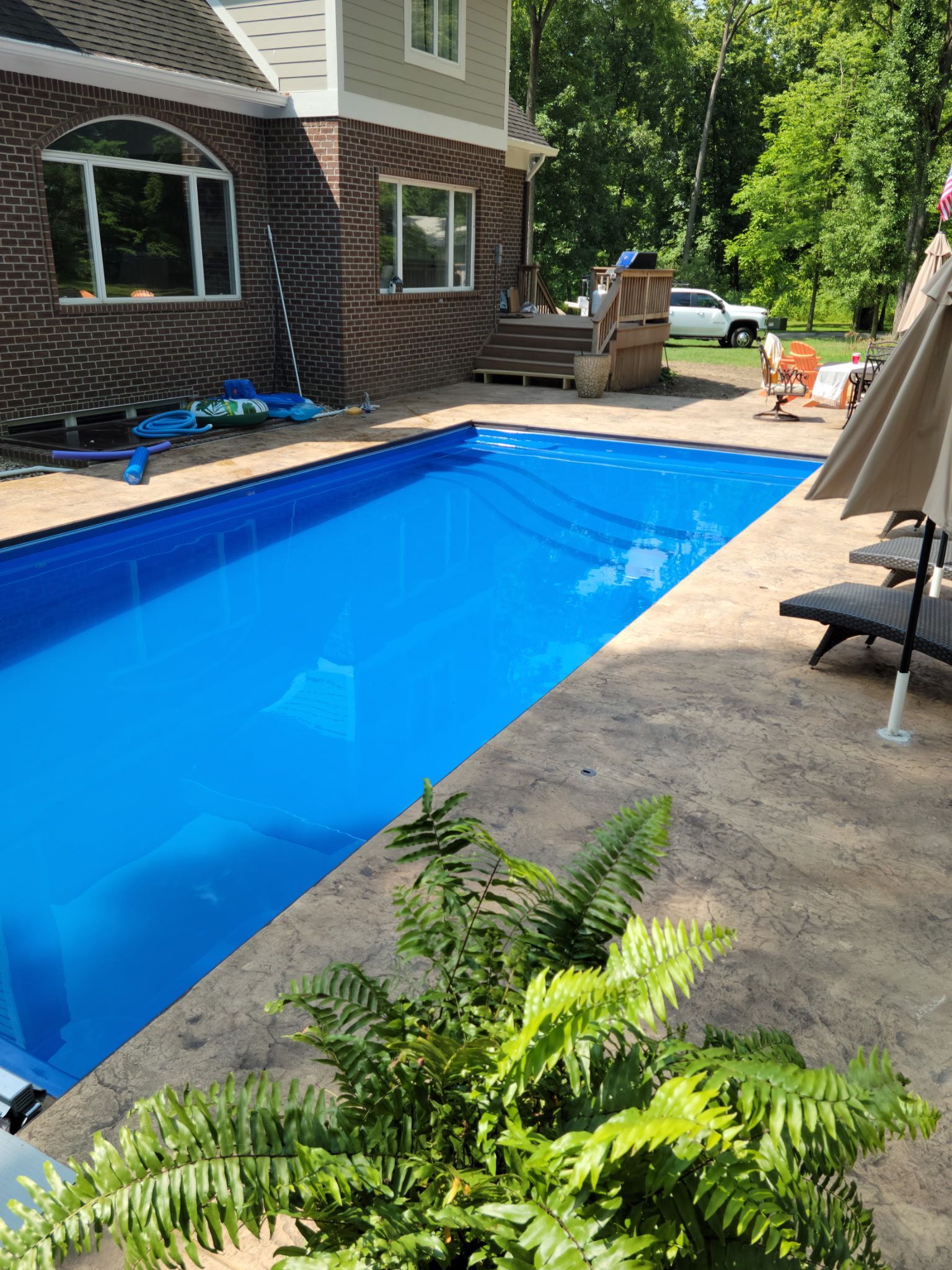 Rectangular pool with blue cover beside a house with brick and tan siding, surrounded by a tan patio.