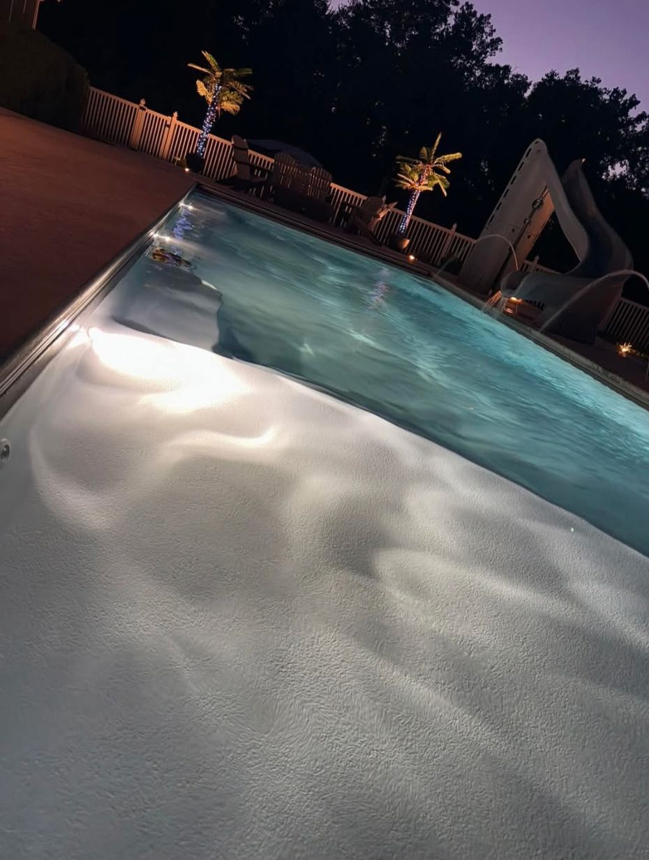 Pool at night, illuminated, with reflected light patterns. Wooden deck and trees in the background.