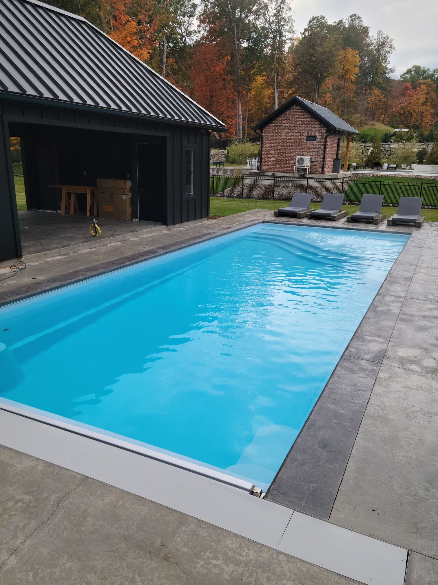 Rectangular swimming pool with blue water, near a dark shed and small stone building; autumn trees in the background.