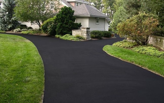 A black driveway leading to a house surrounded by grass and trees.