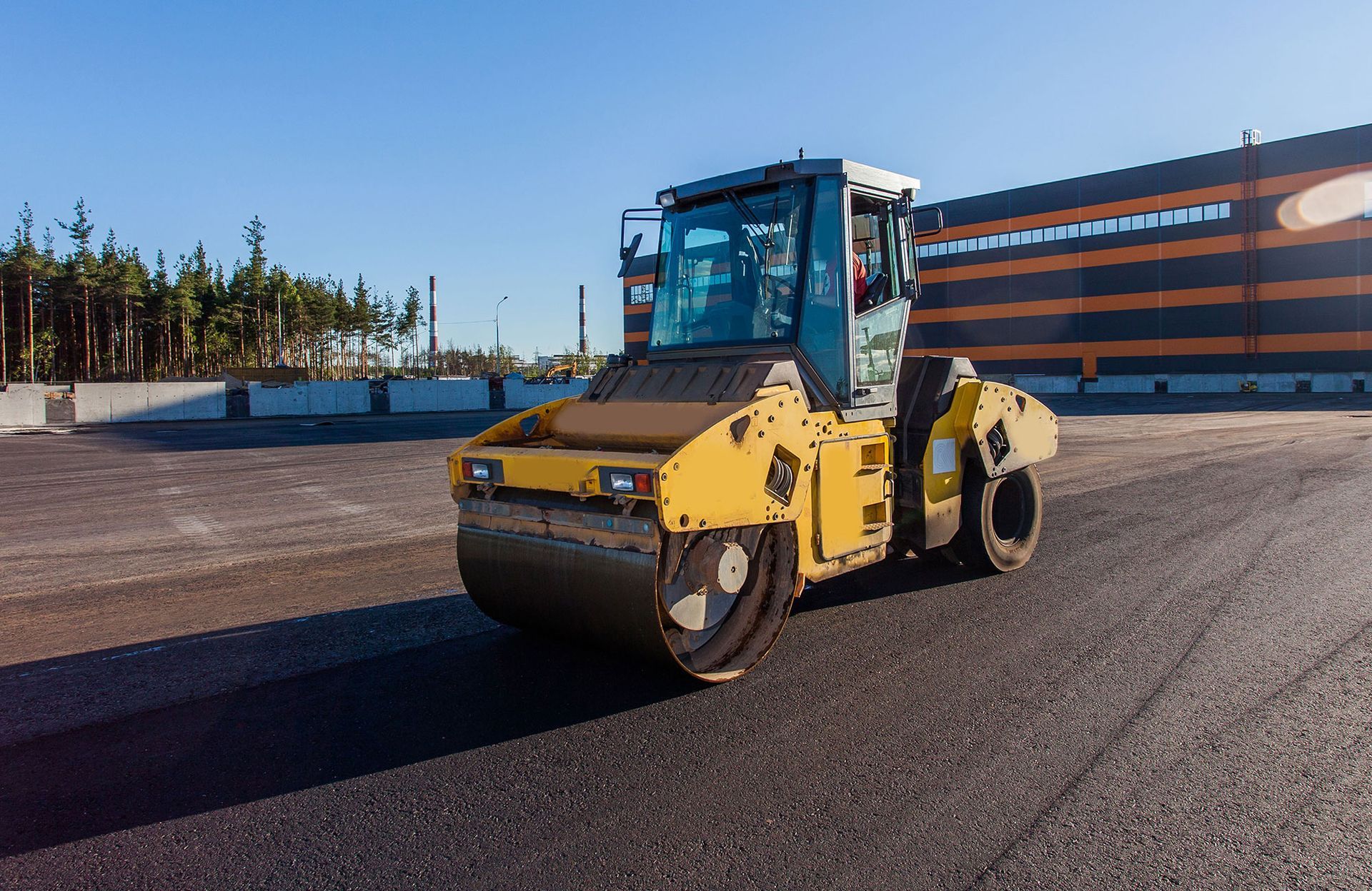 A yellow roller is sitting on top of a dirt road in front of a building.
