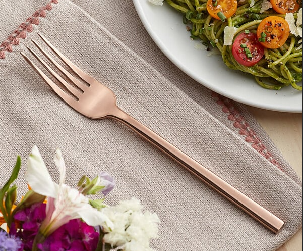 Rose gold fork on a linen napkin next to a plate of pasta and flowers.