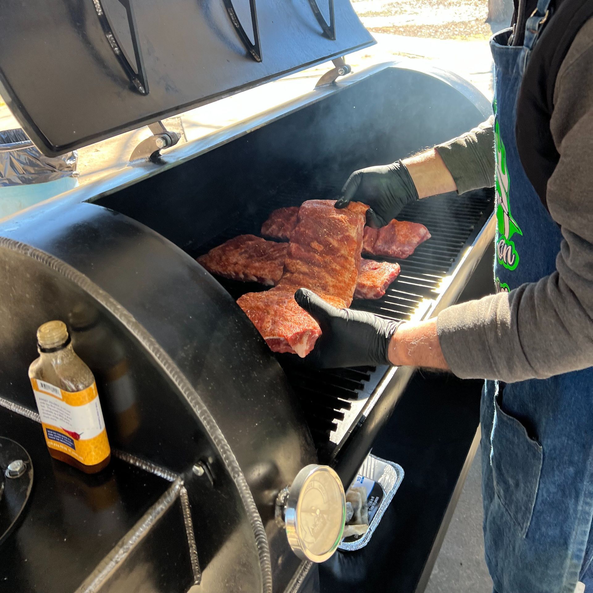 A man is cooking meat on a grill with a bottle of bbq sauce in the background