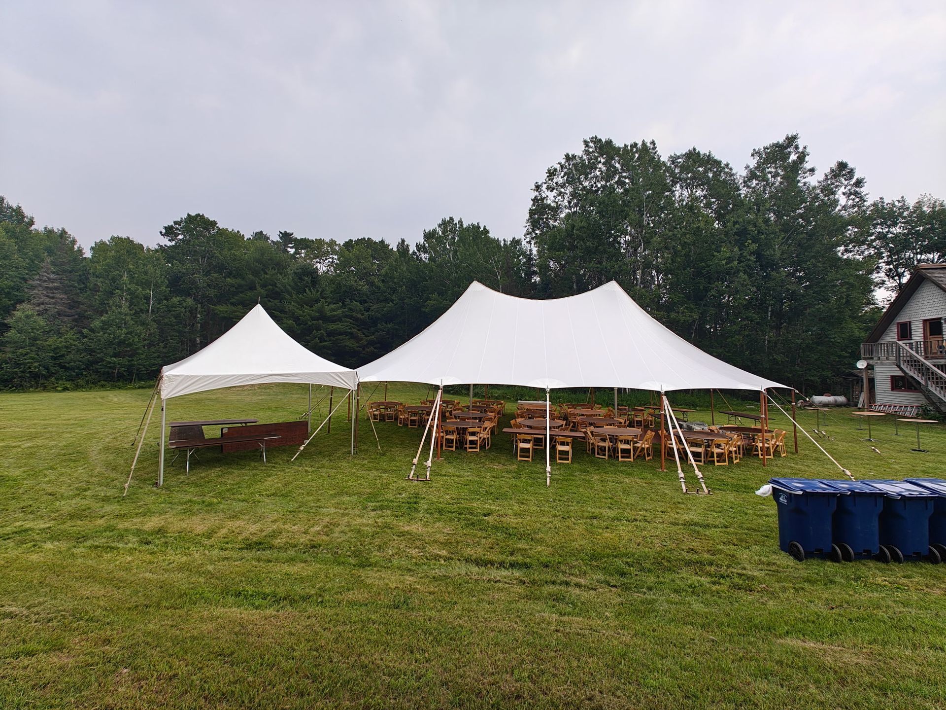 Two white tents in a grassy field with tables and chairs set up, and trees in the background.