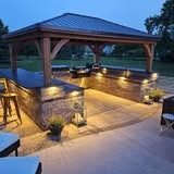 Outdoor kitchen with wooden gazebo and built-in grill, illuminated by warm lights at dusk.