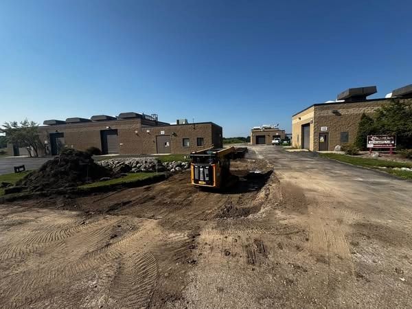 A small construction vehicle excavating a ditch between two brick buildings under a blue sky.