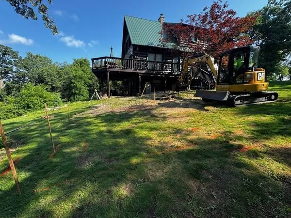 Construction site with excavator near a wooden house on a grassy hillside.