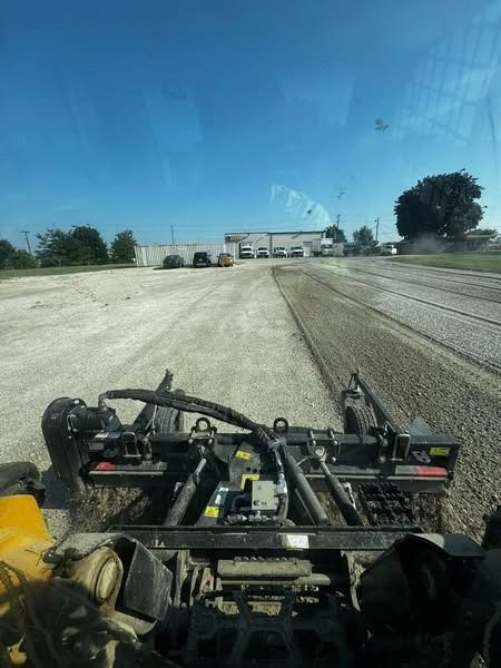 A road grader leveling gravel on a sunny day. Buildings and trees in the background.