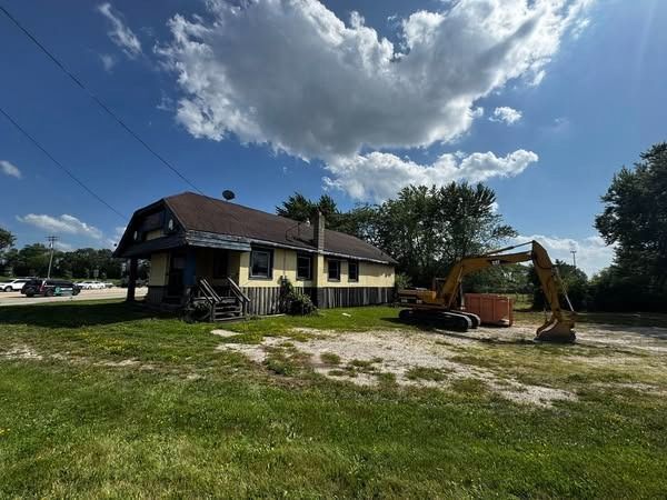 An excavator next to a yellow building under a partly cloudy sky, with grassy foreground.