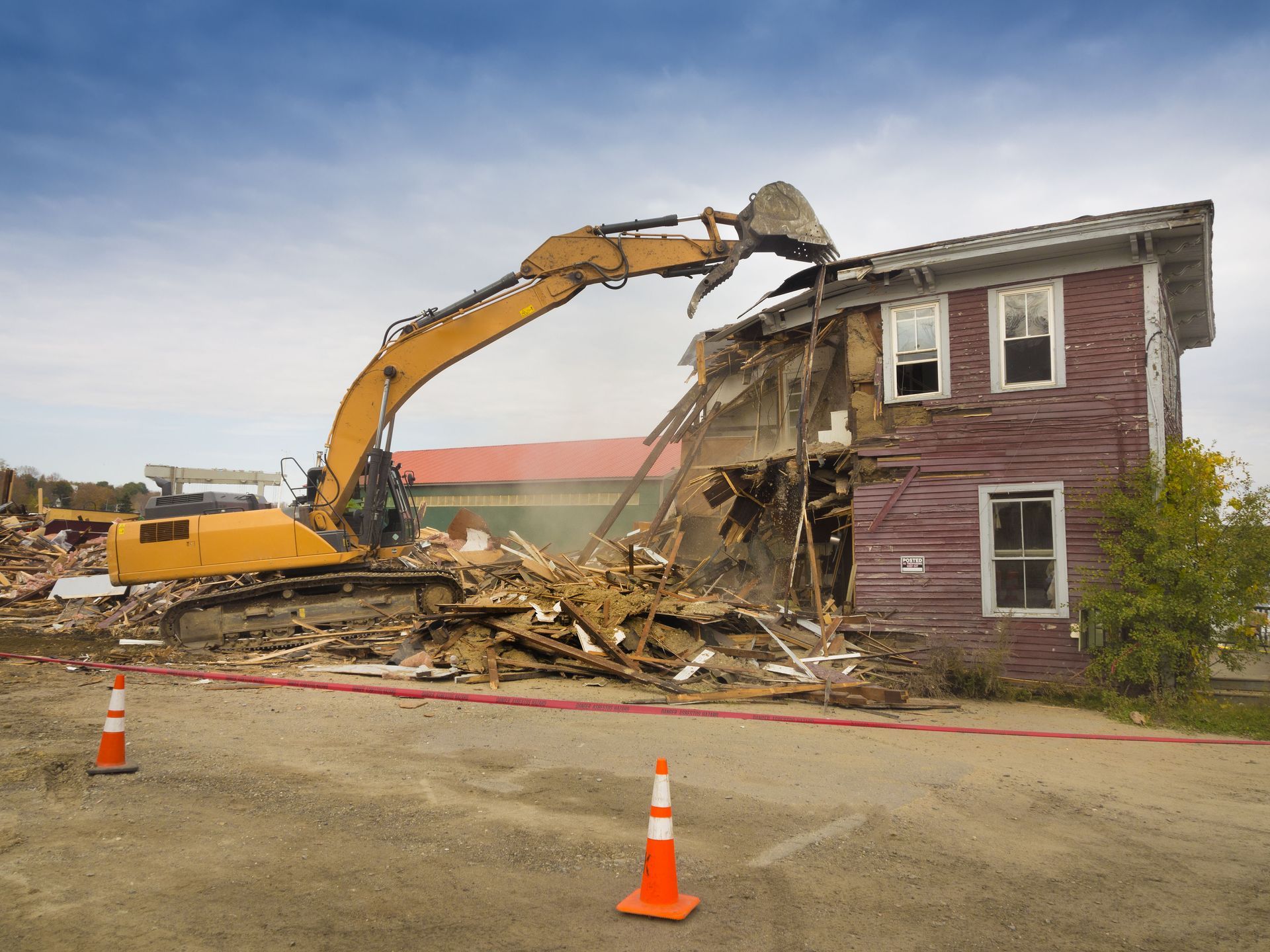 A yellow excavator demolishes a red wooden building, debris scattered, orange safety cones in foreground.