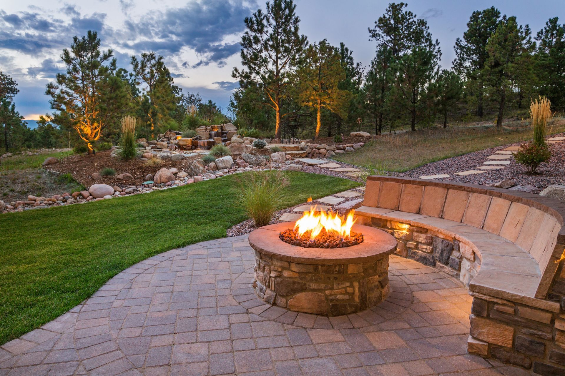 Fire pit with flames, stone bench, paved patio. Scenic outdoor setting with grass and trees at dusk.
