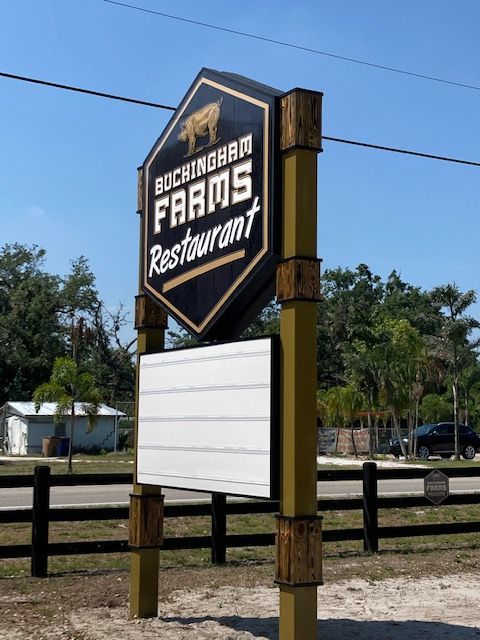 A roadside sign for Buckingham Farms Restaurant, featuring a hexagonal black logo on yellow posts with a blank message board.