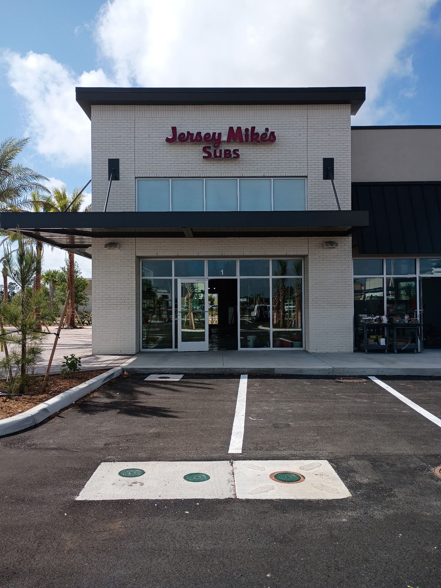A storefront for a Jersey Mike's Subs restaurant on a sunny day, featuring a modern exterior and an adjacent parking lot.
