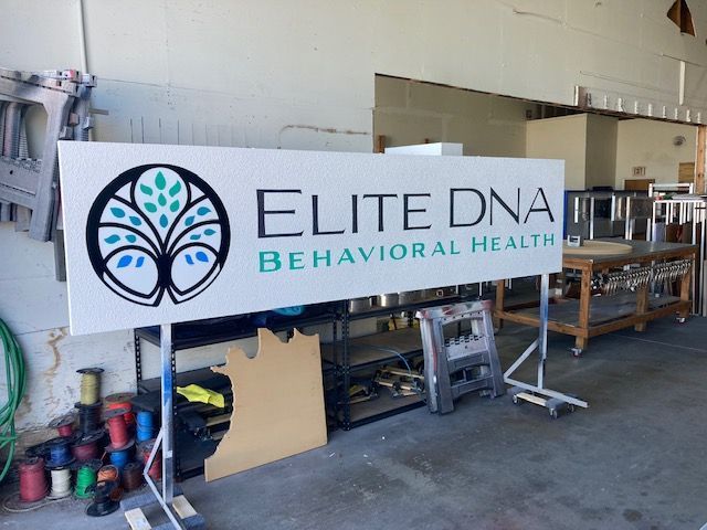 A white sign for Elite DNA Behavioral Health featuring a tree logo with blue and green leaves, standing in a workshop.