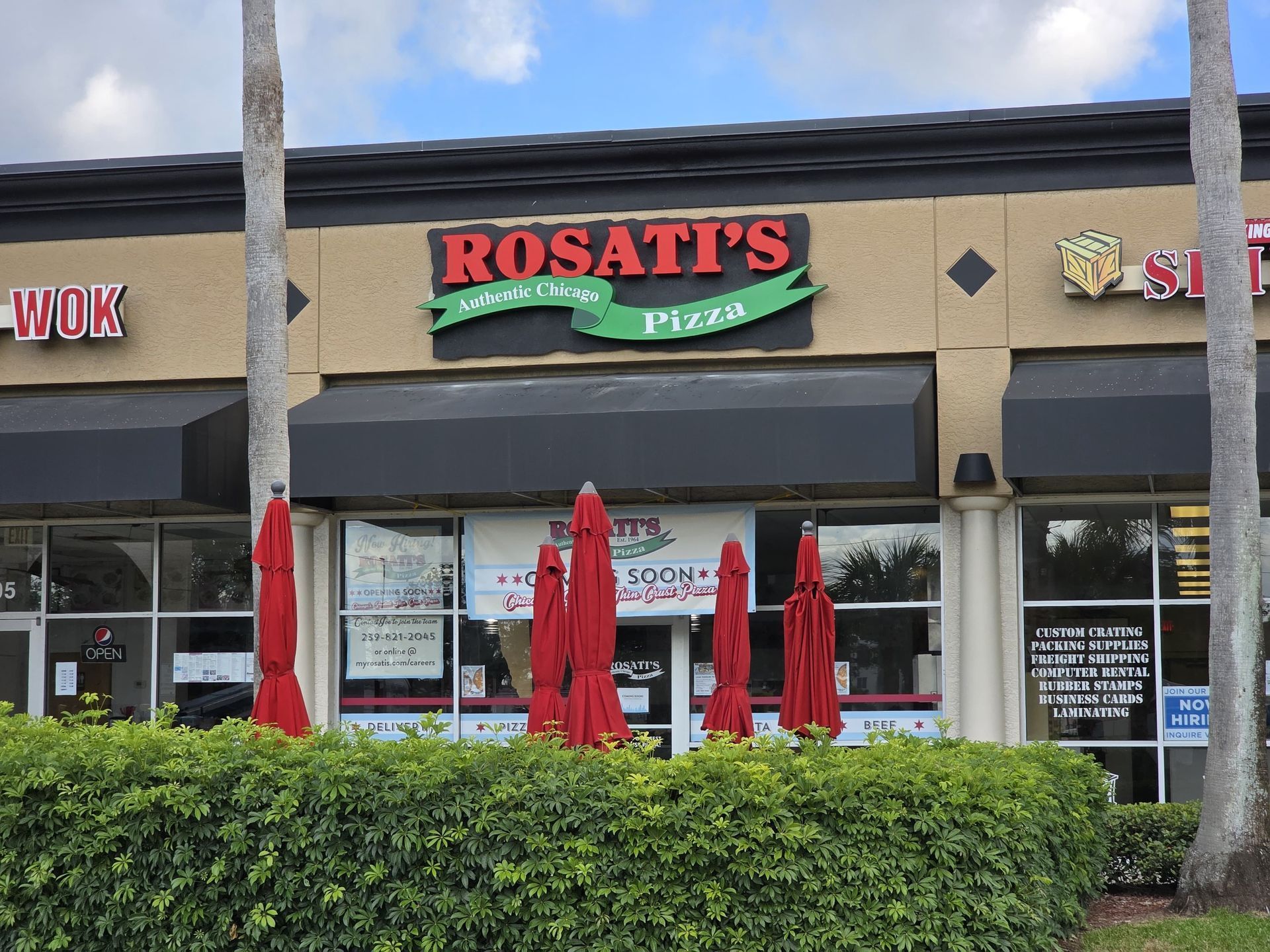 Rosati's Pizza restaurant storefront in a strip mall with red umbrellas and green hedges in front.