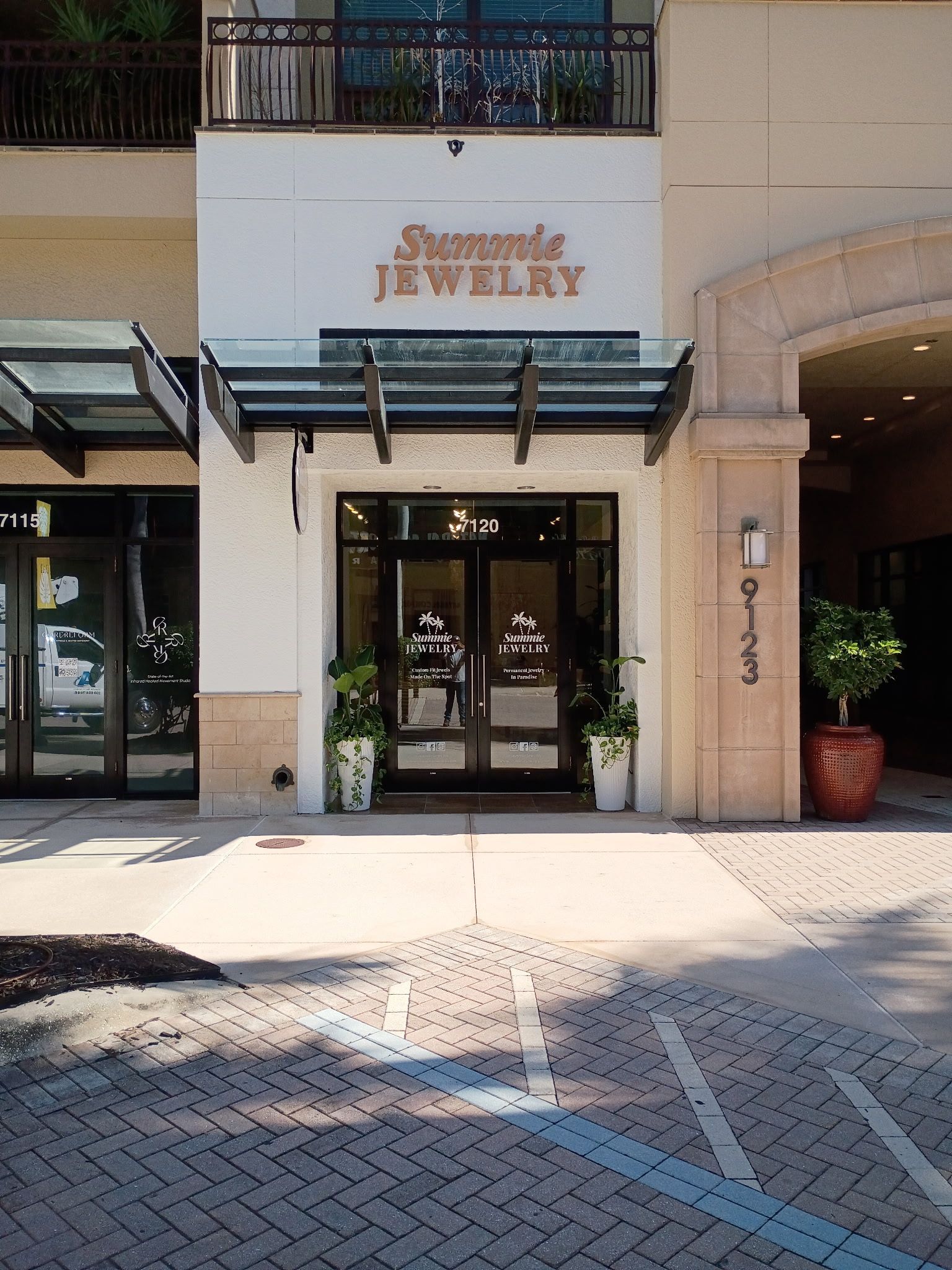 The storefront entrance of Simone Jewelry, featuring glass double doors, an awning, and potted plants on a brick sidewalk.