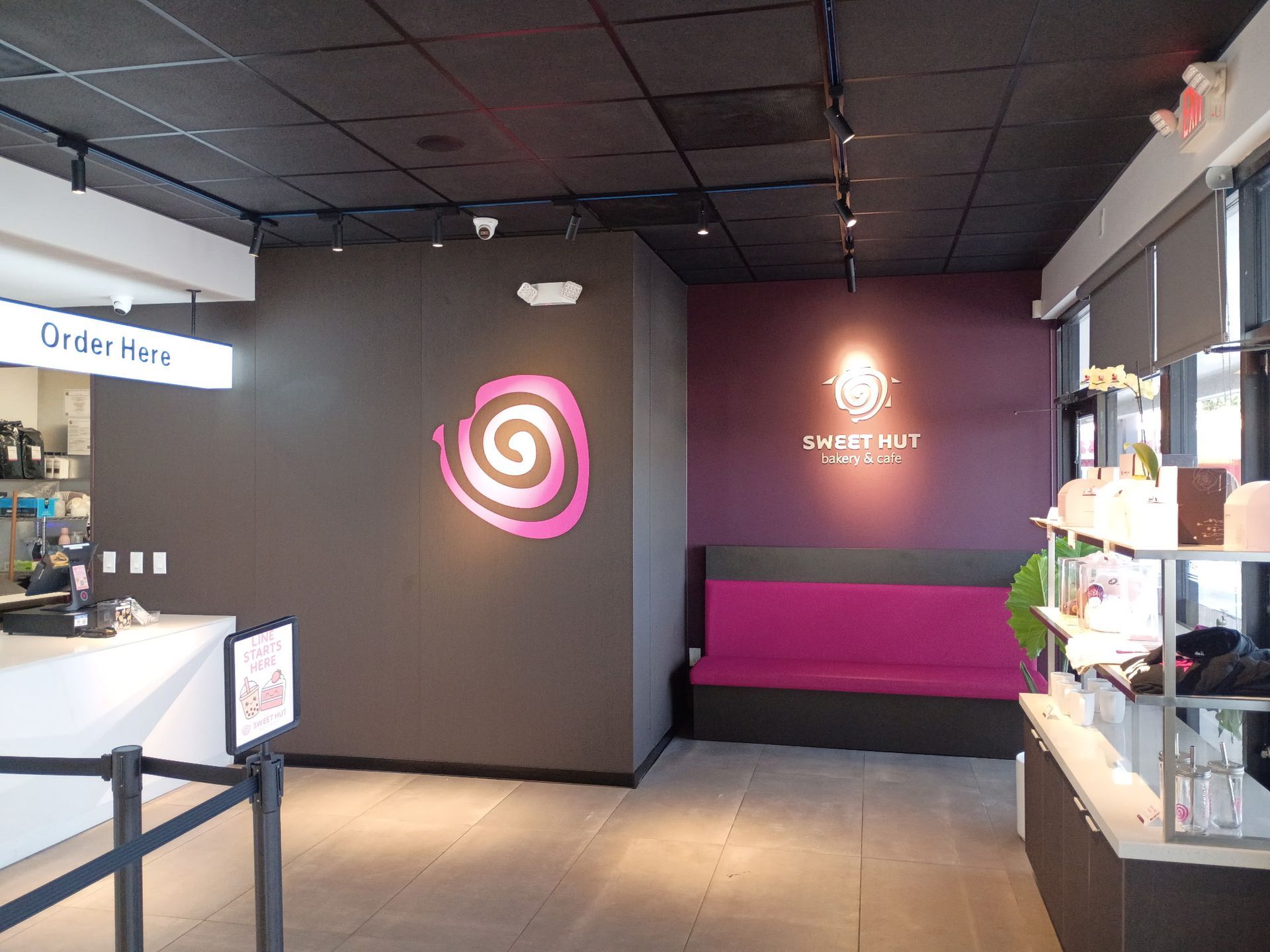 The interior of Sweet Hut Bakery & Cafe with a counter, a purple bench, and a spiral pink logo on a dark wall.