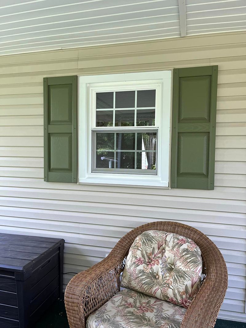 A wicker chair is sitting on a porch next to a window with green shutters.
