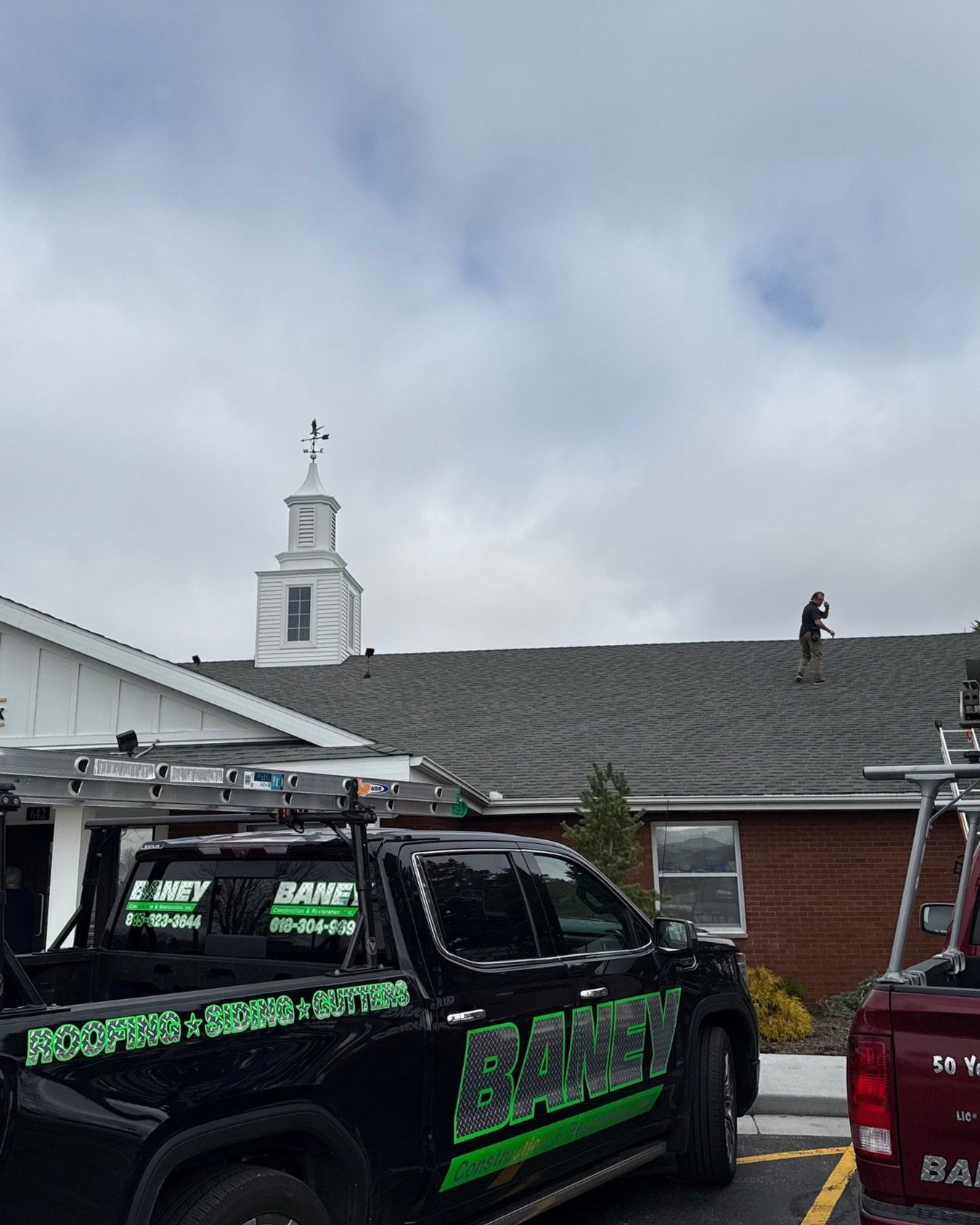 Damaged house roof with a worker on top and utility trucks parked below under a cloudy sky
