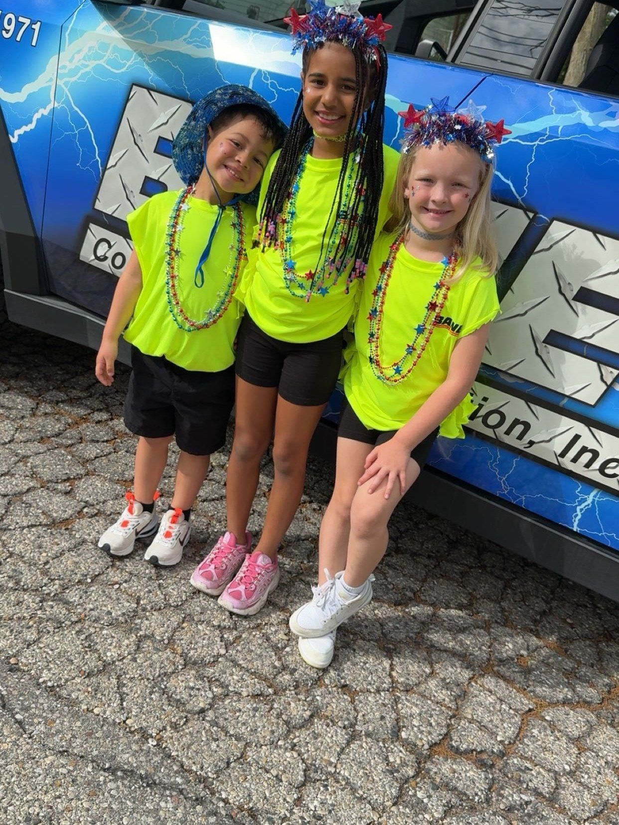 Three children in neon shirts and star headbands pose in front of a decorated vehicle.