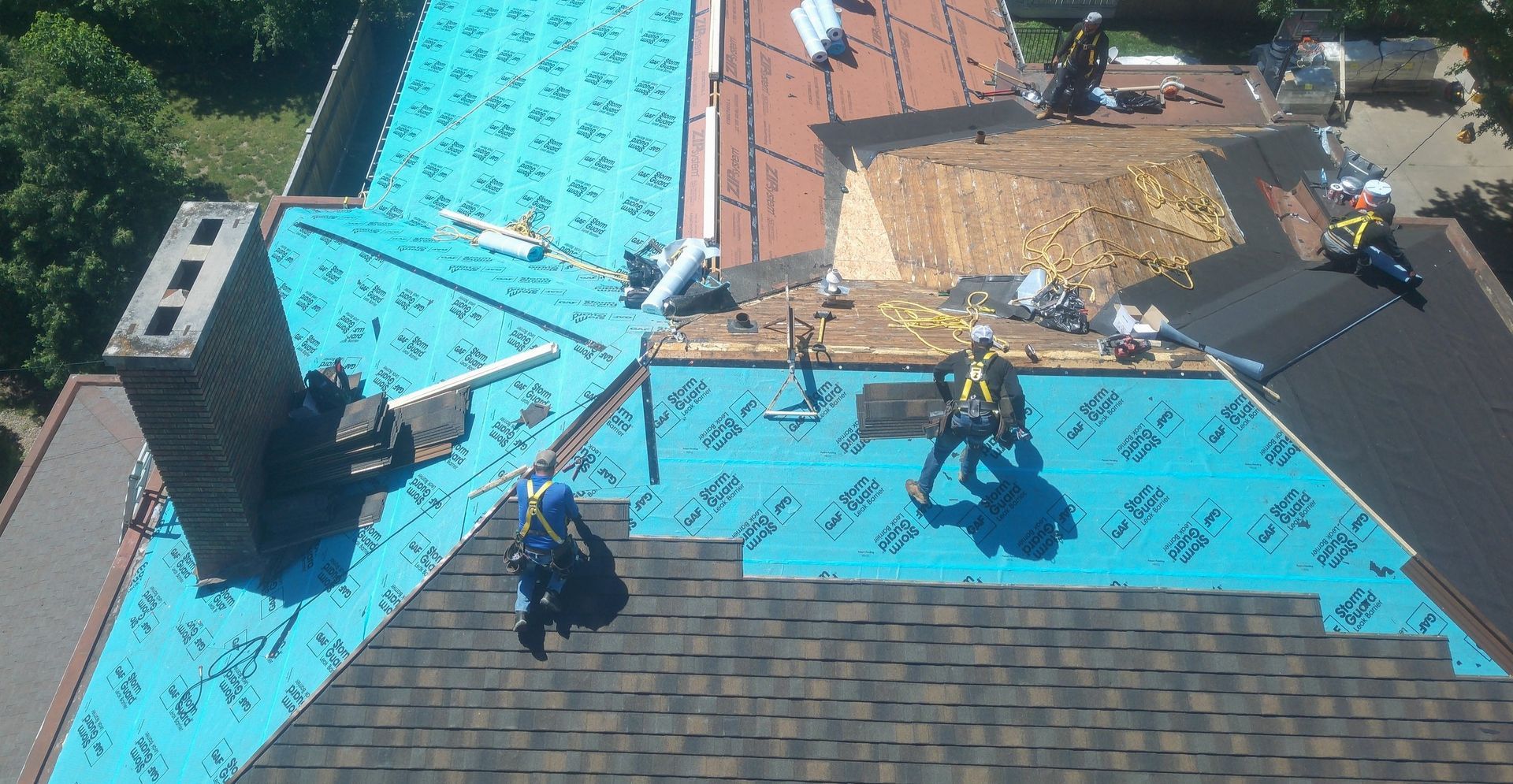 Workers installing bright blue roofing on a house with piles of materials and tools visible nearby