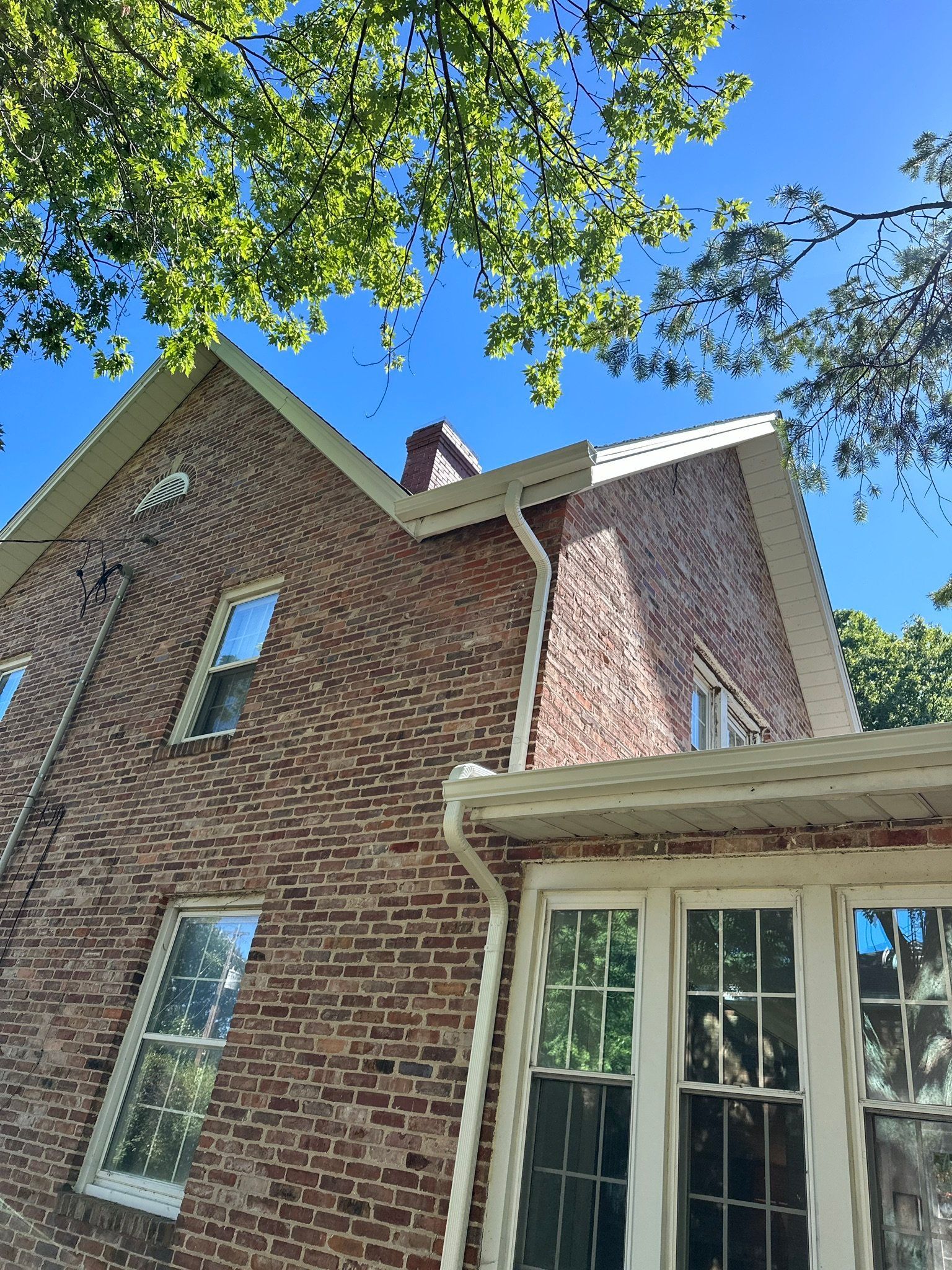 Brick house exterior with tall windows, white trim, and blue sky framed by green tree branches