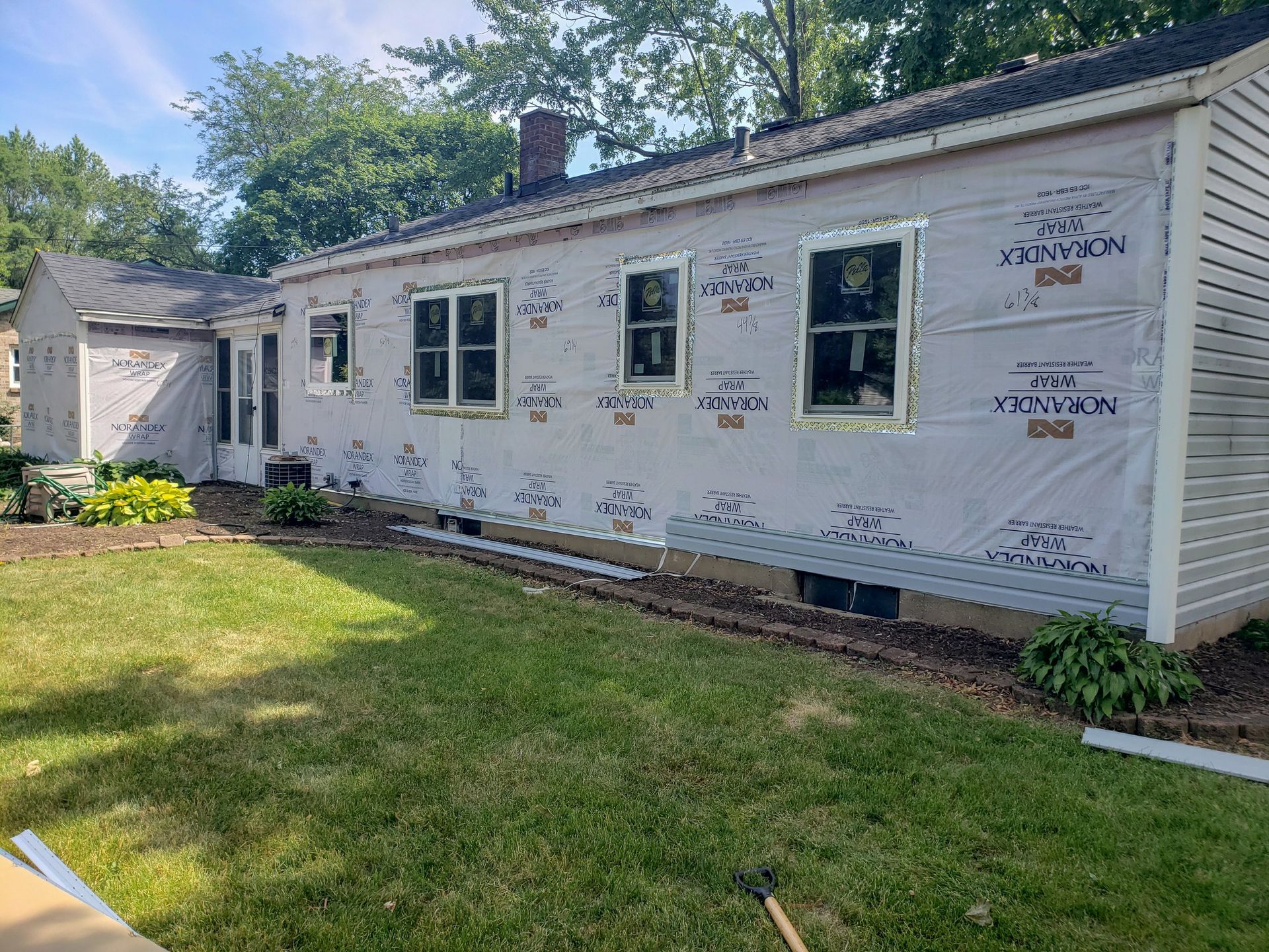 Single-story house under construction with white house wrap, new windows, and a green lawn in front