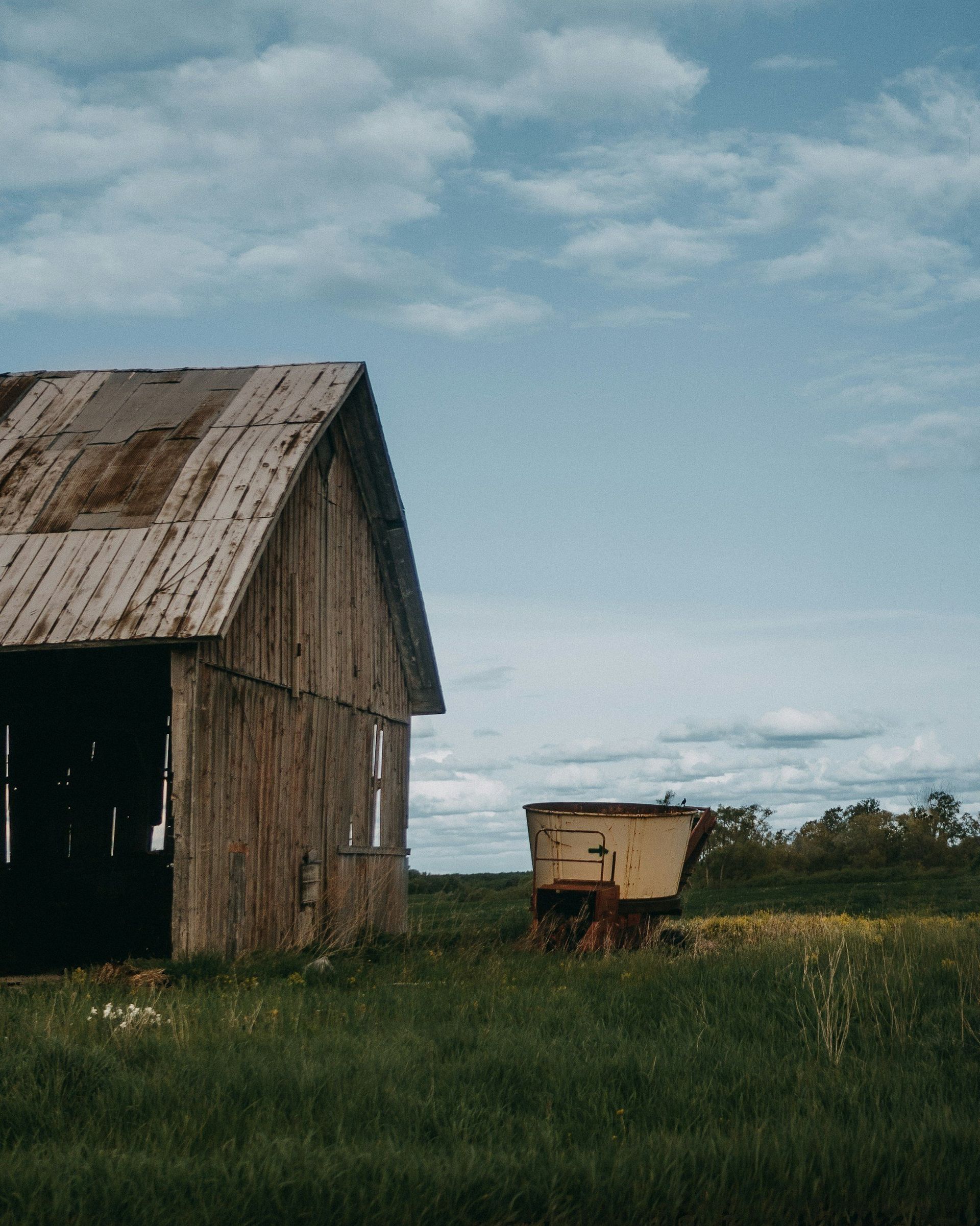 Weathered wooden barn beside a small cart in a grassy field under a cloudy blue sky