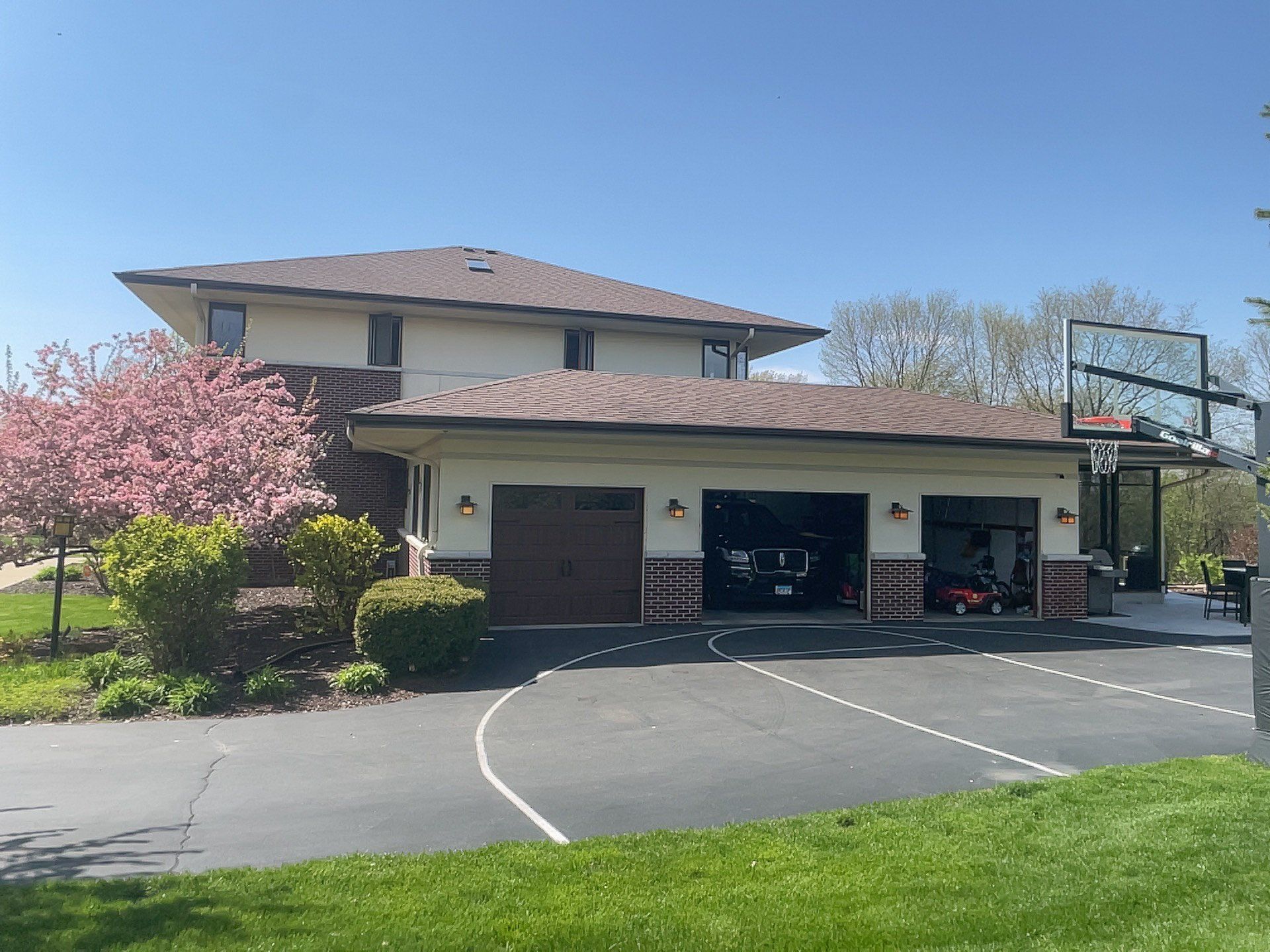 Suburban house with attached garage and driveway, blooming pink tree, and a basketball hoop under a blue sky