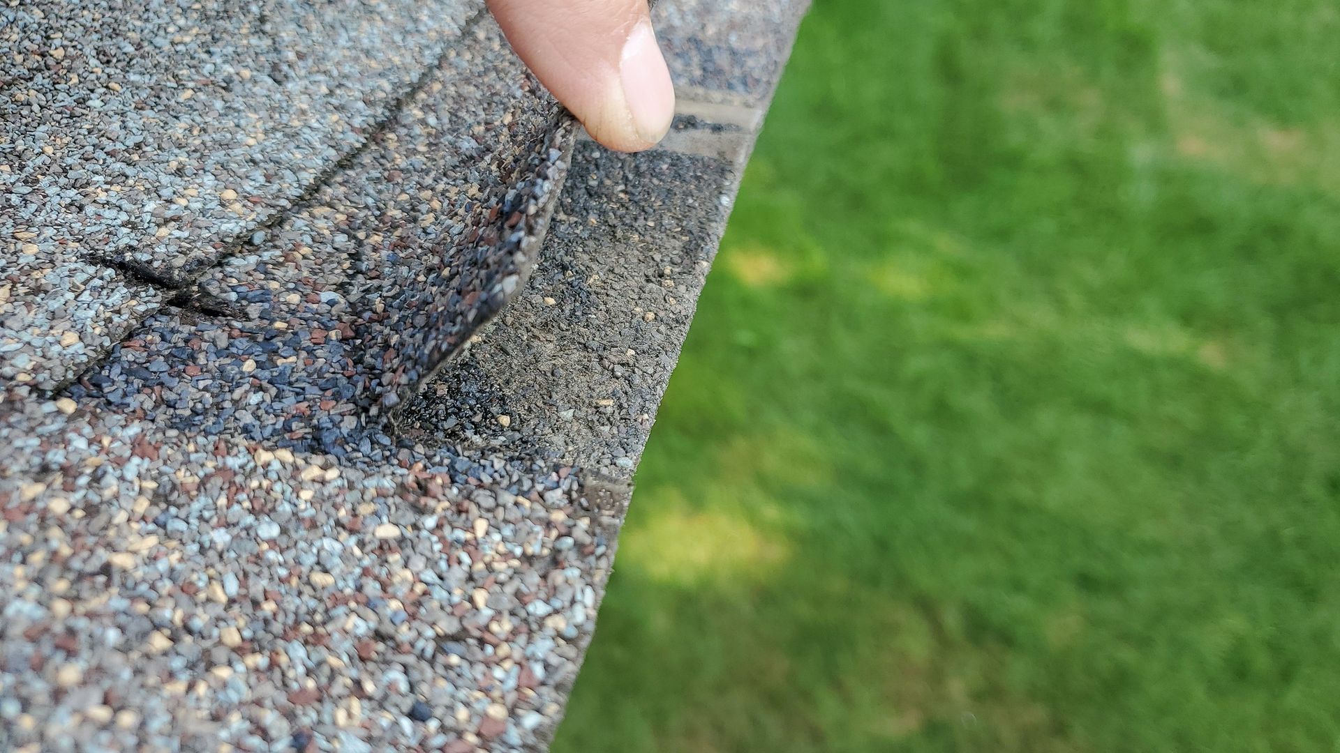 Hand pressing up a curled edge of weathered roof shingles beside a grassy lawn