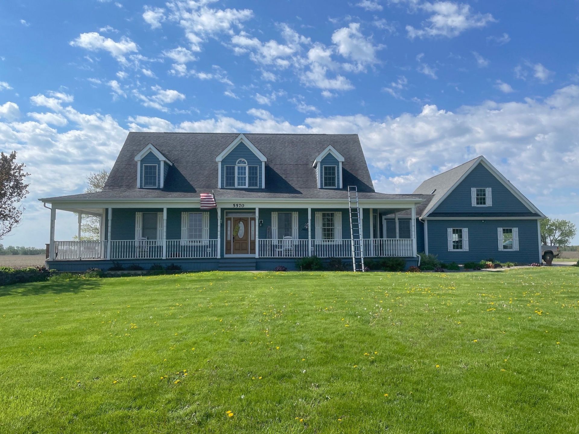 Blue farmhouse with white porch and dormer windows under a partly cloudy sky, set behind a green lawn
